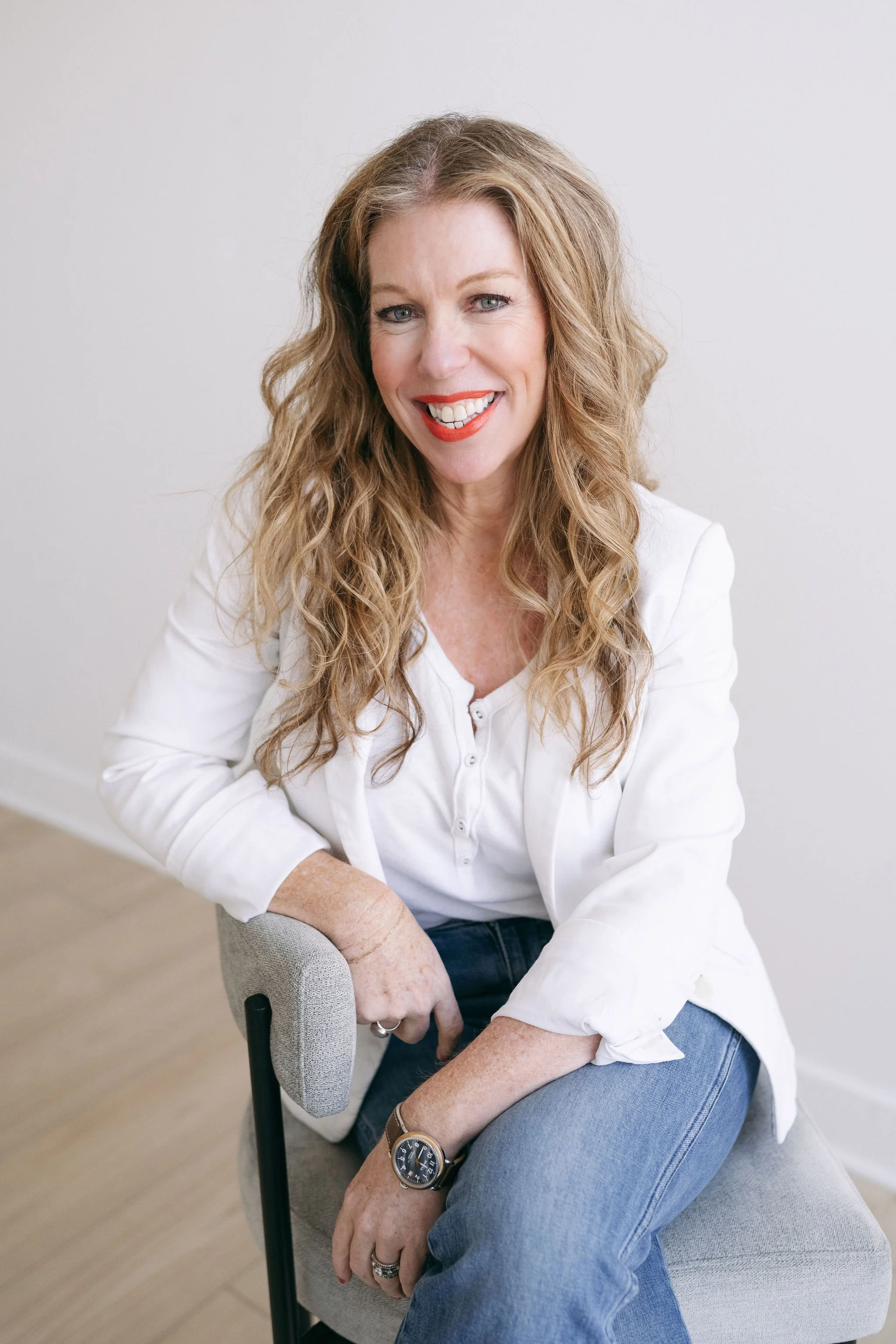 A woman with long, curly hair, wearing a white shirt and blue jeans, sitting and smiling at the camera.