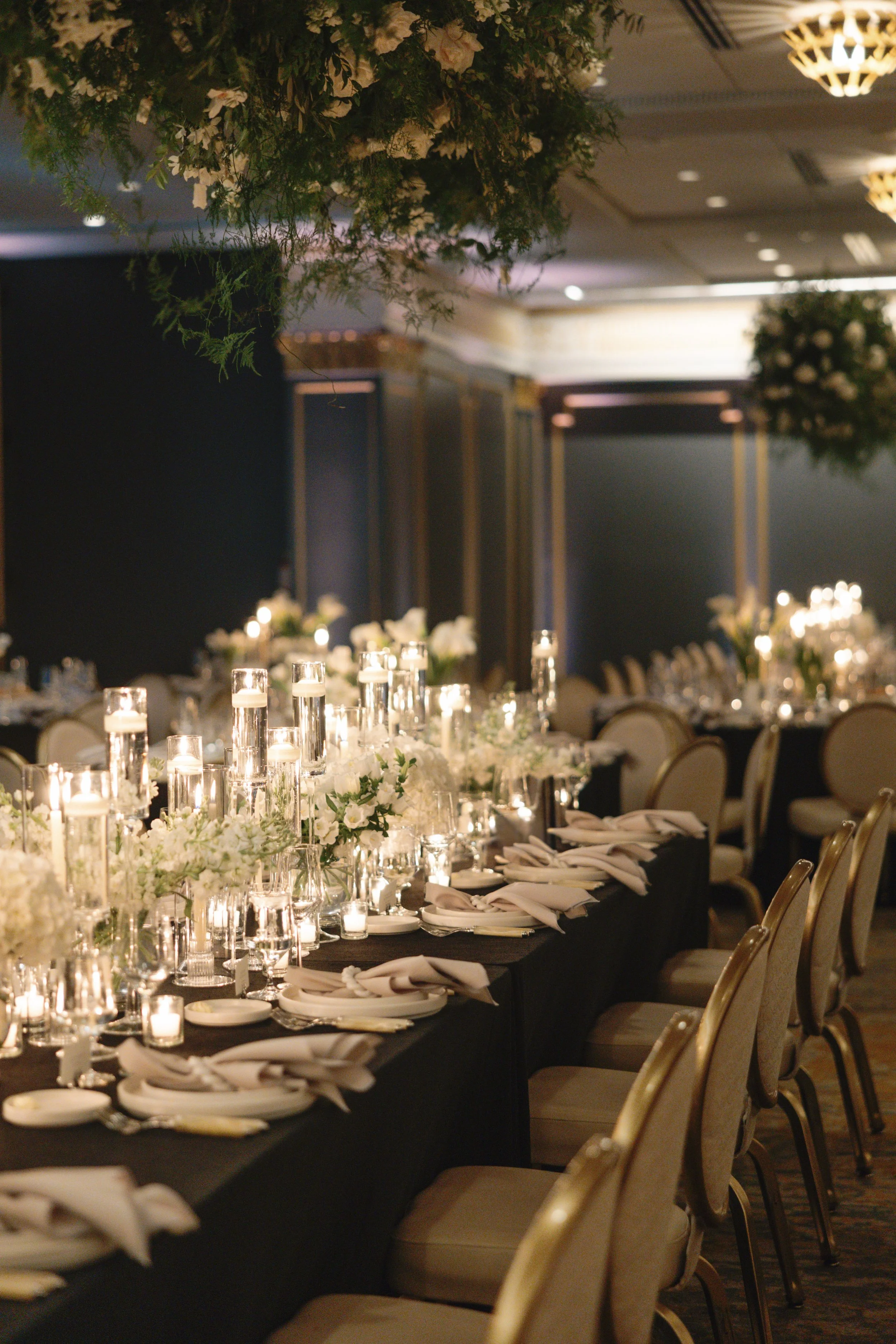 Elegant event banquet table decorated with white flowers, candles in glass holders, and neatly folded napkins at each place setting in a dimly lit room.