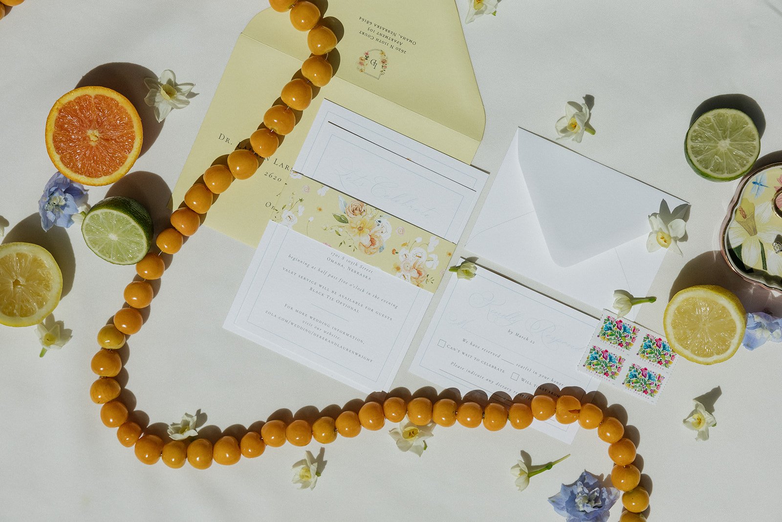 Flat lay of wedding invitation cards, yellow beads, and slices of citrus fruits including orange, lime, and lemon on a white surface, decorated with small white and blue flowers.