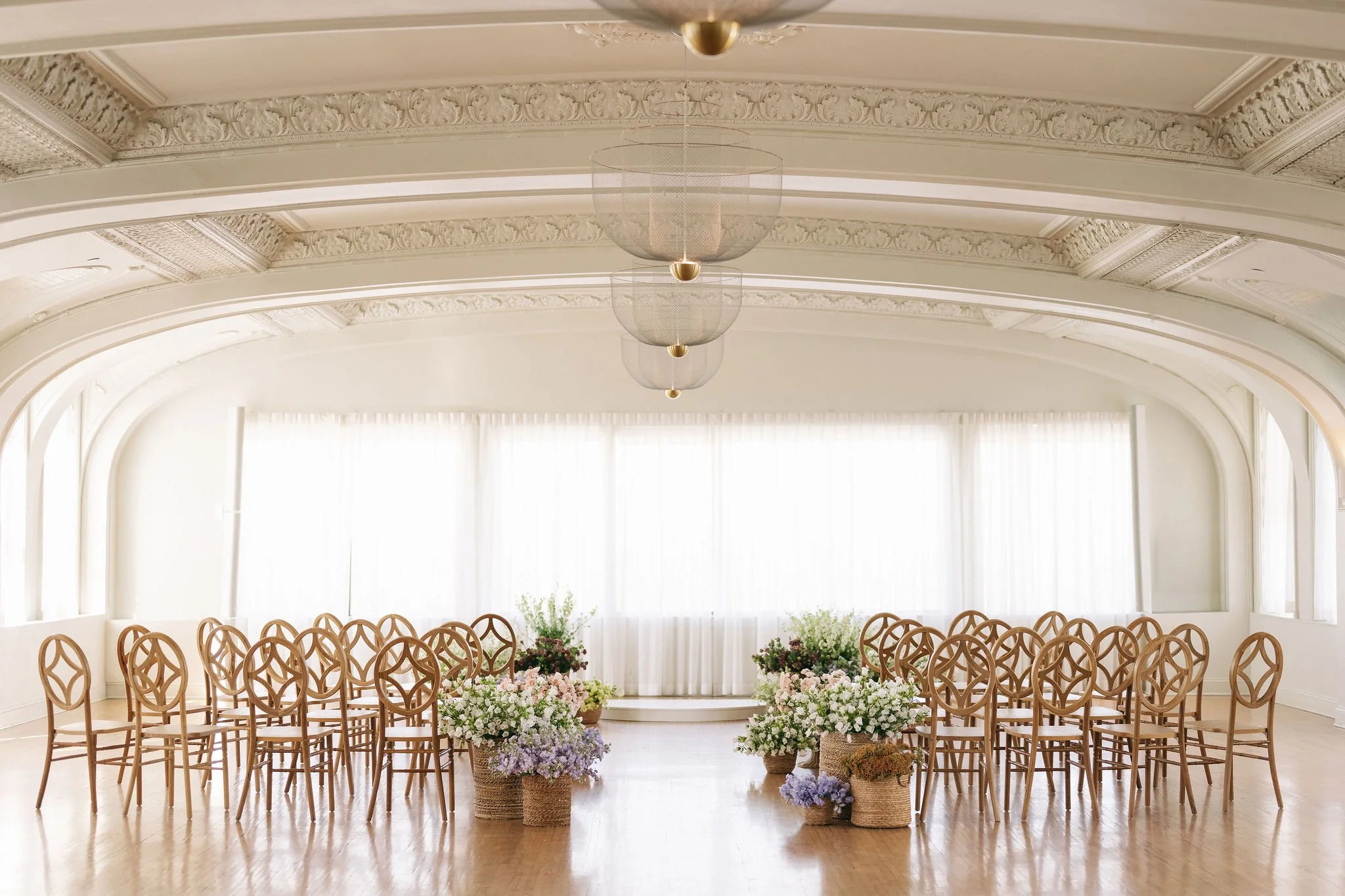 Empty wedding or event ceremony room with wooden chairs arranged in rows, floral arrangements in baskets at the front, and large windows with white curtains, with intricate ceiling moldings and three hanging light fixtures.