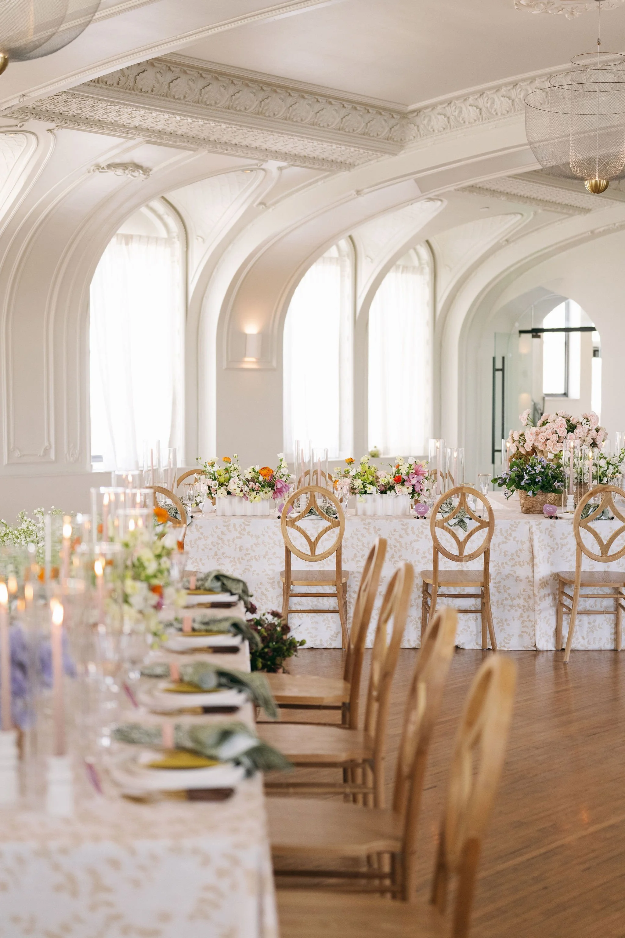 Elegant banquet hall decorated for a celebration with a long table, floral arrangements, candles, and chairs with intricate backs, set in a bright room with arched windows and ornate ceiling details.