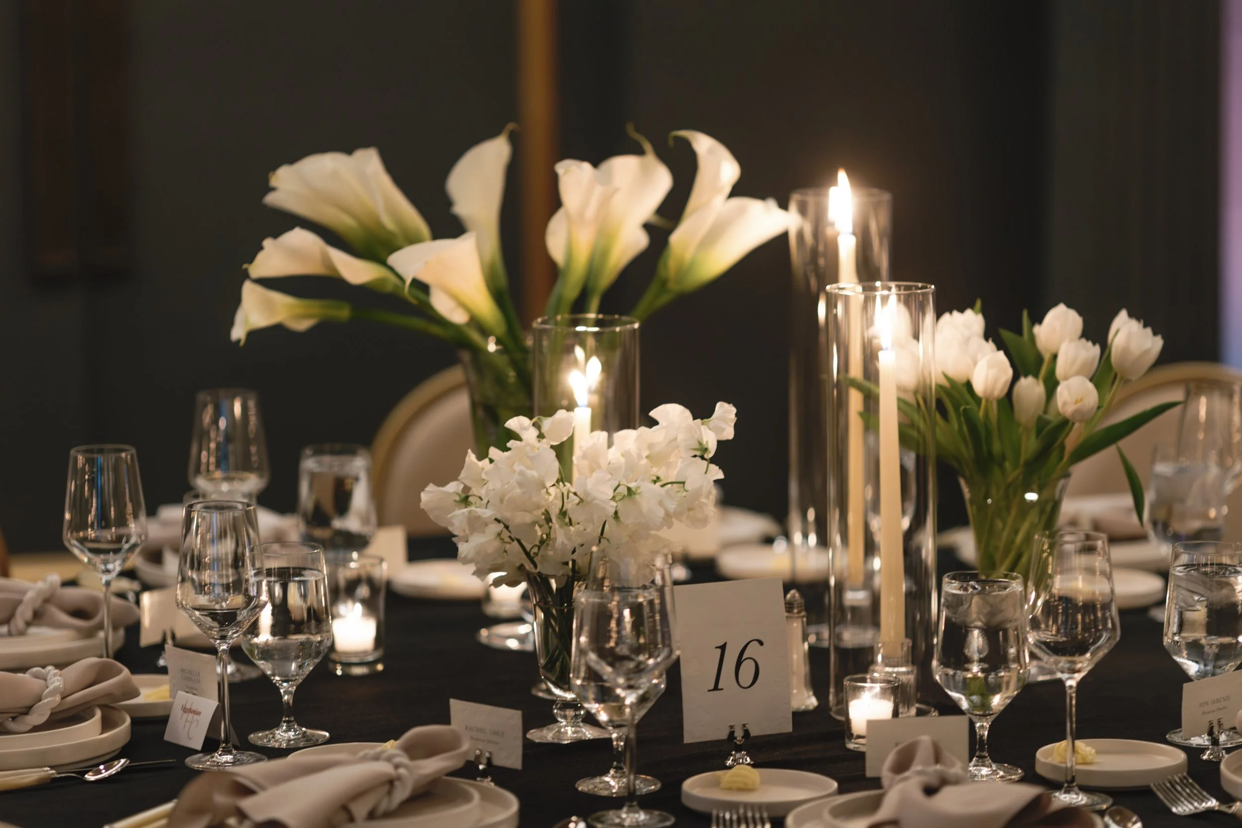 Elegant dinner table decorated with white flowers, tall candles in glass holders, black tablecloth, white plates, silverware, and clear glassware, with a table number 16 displayed.