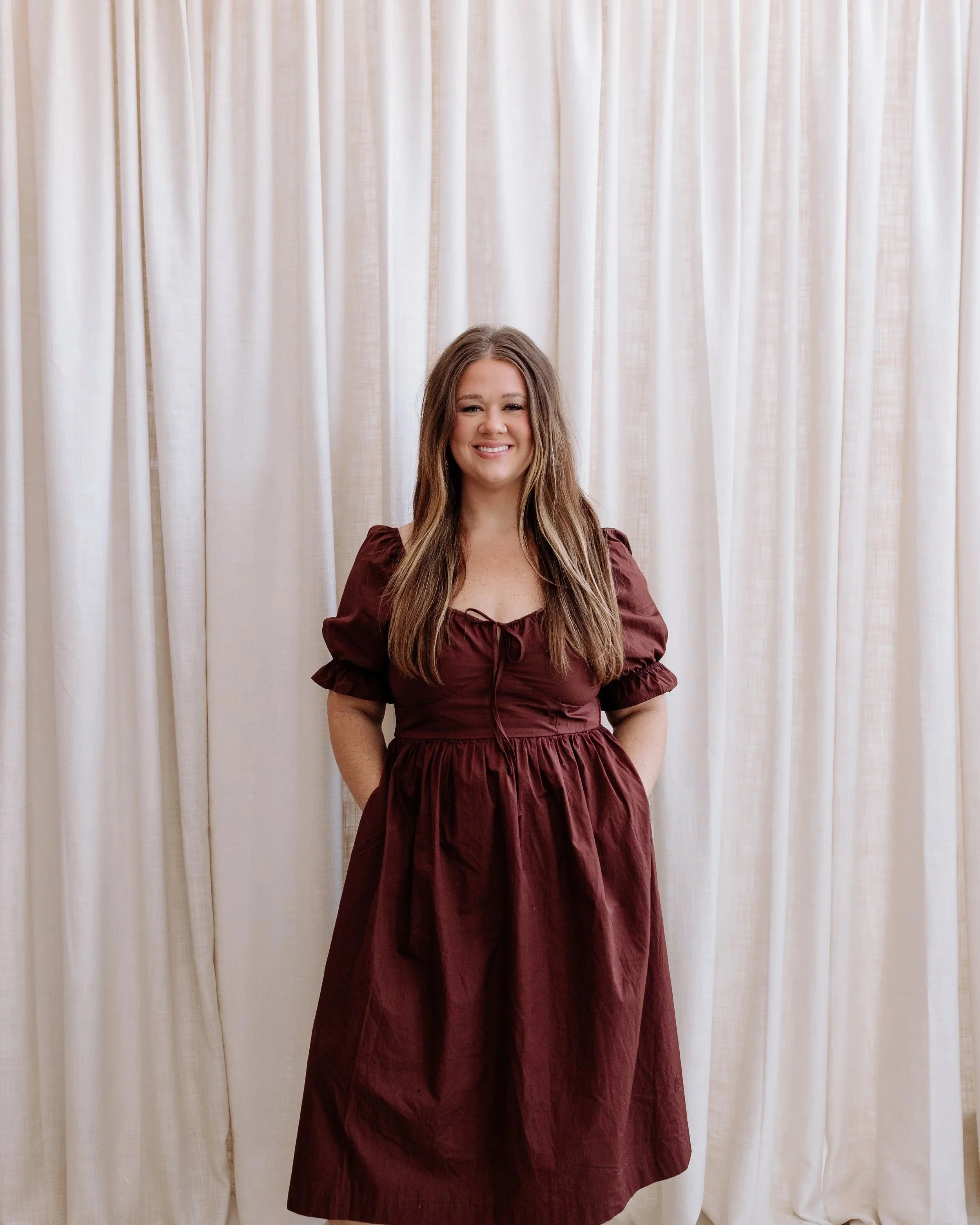 A woman with long, curly brown hair smiling and looking at the camera against a plain white background.
