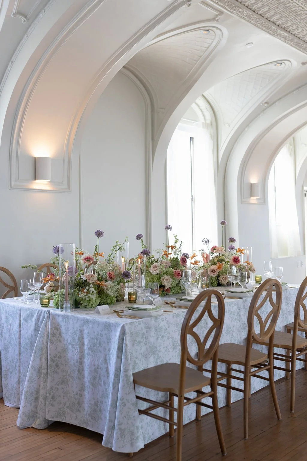 Elegant indoor dining table decorated with pastel flowers and tall candles, set for a formal event in a brightly lit room with arched windows and ornate white walls.