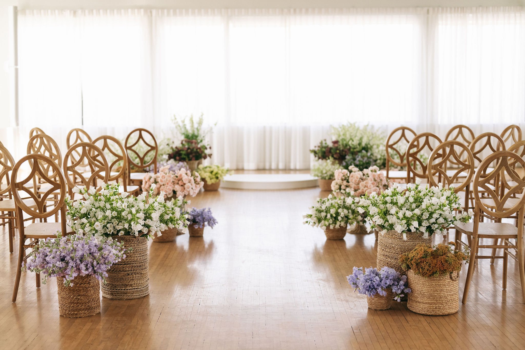 Empty wedding or event ceremony space with rows of wooden chairs and flower arrangements in woven baskets along the aisle, in front of a bright windowed backdrop.