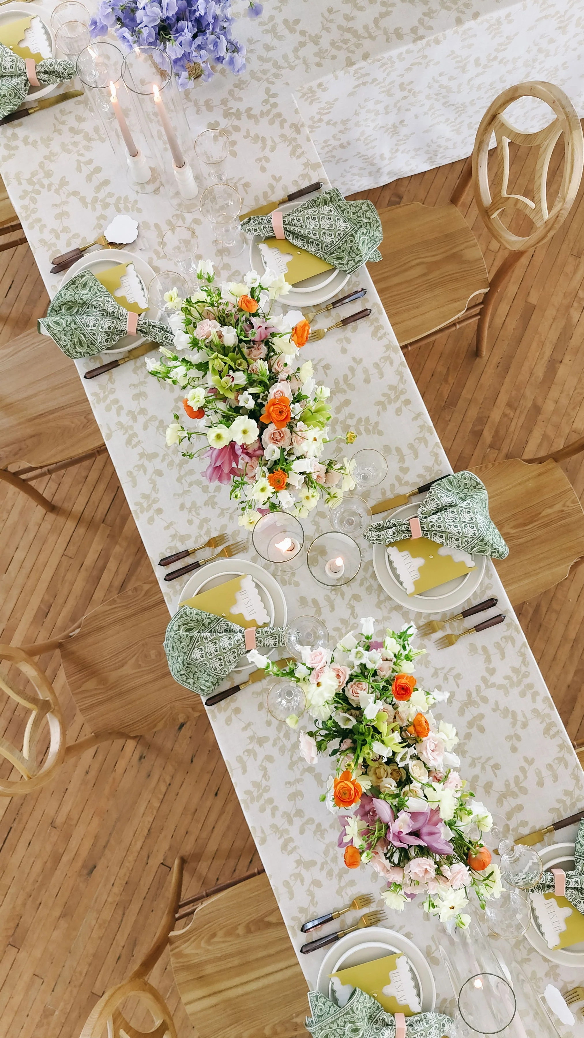 A long dining table set for a celebration with floral centerpieces, candles, and tableware, viewed from above.