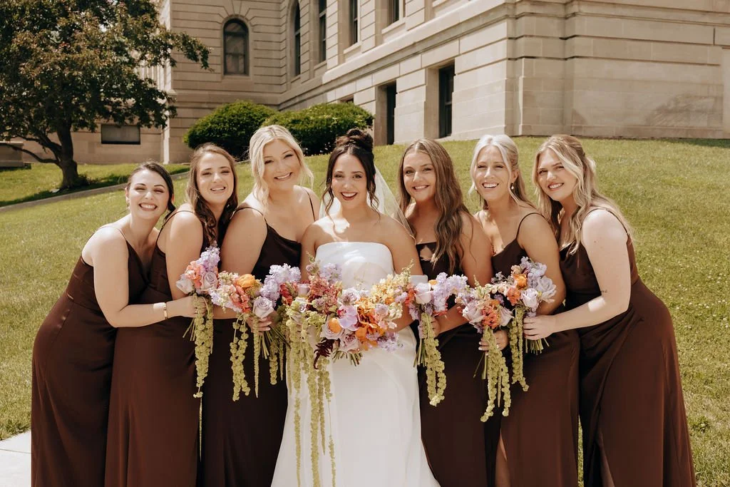 A bride in a white dress standing with six bridesmaids in dark brown dresses, holding bouquets of pink, purple, and orange flowers outside a stone building with green grass and trees.