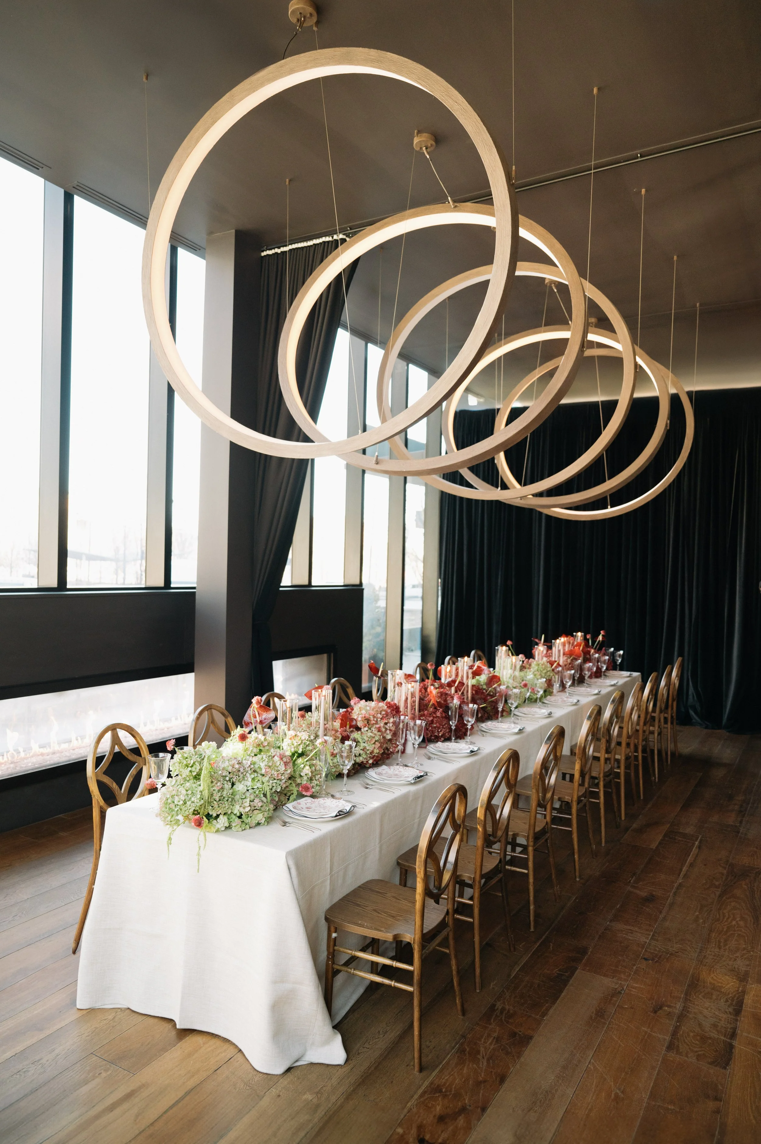 Elegant dining room with a long table set for an event, decorated with floral centerpieces, wine glasses, and cutlery. Overhead, modern circular light fixtures hang from the ceiling. The room features large windows with black curtains, and wooden flo