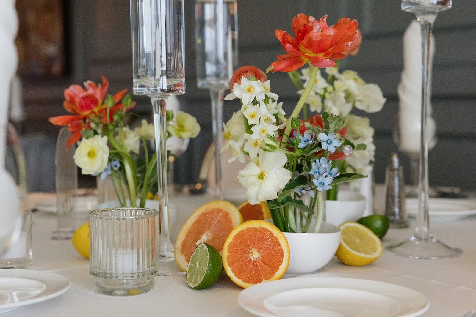 A table centerpiece with fresh flowers in a white bowl, surrounded by halved lemons and limes, and empty wine glasses.