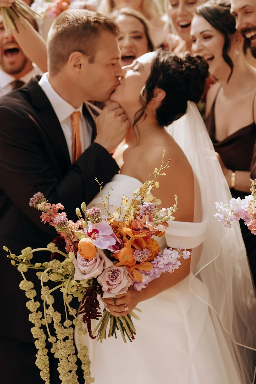 A bride and groom share a kiss at their wedding, surrounded by friends. The bride holds a bouquet of pink, purple, and orange flowers.
