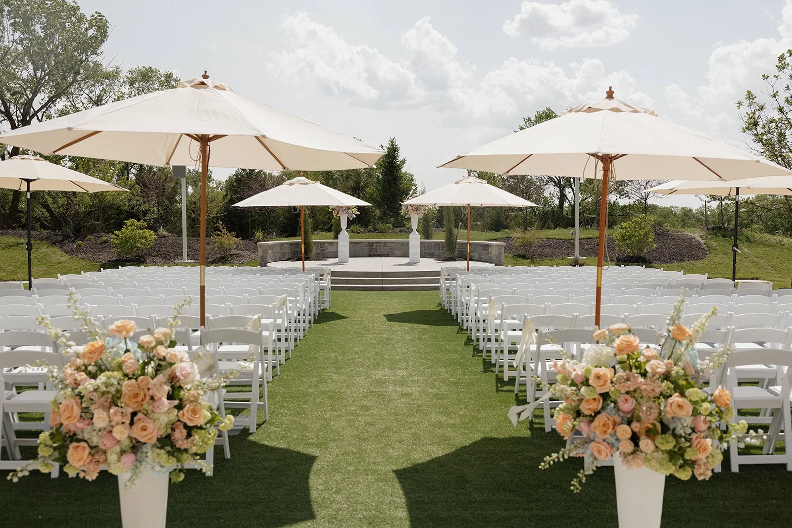 Outdoor wedding setup with white chairs arranged in rows on a grassy area, decorated with large floral bouquets in white vases at the entrance, and white umbrellas providing shade, with an outdoor stage and cloudy sky in the background.
