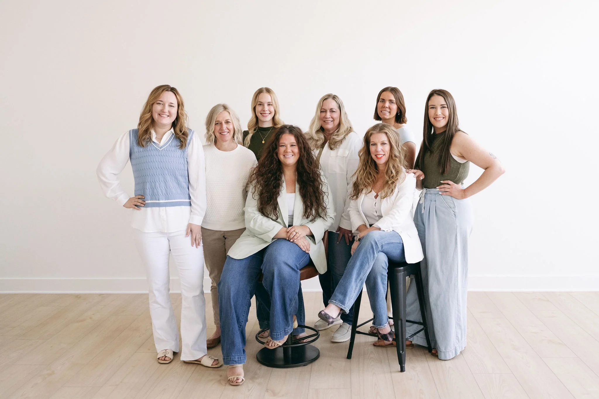 Group of nine women posing together in front of a plain white wall, dressed in casual and business casual clothing.