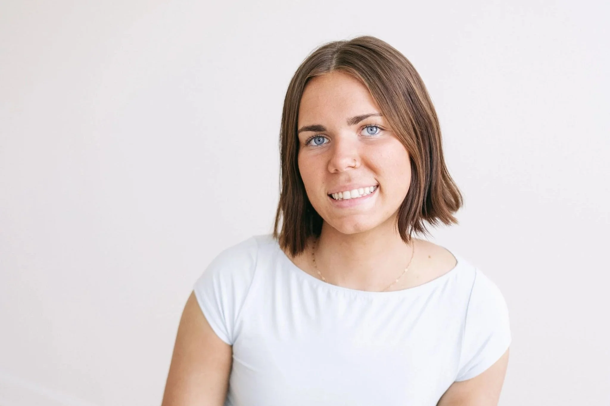 A young woman with short brown hair, blue eyes, and a nose piercing, smiling at the camera, wearing a white t-shirt, against a plain white background.