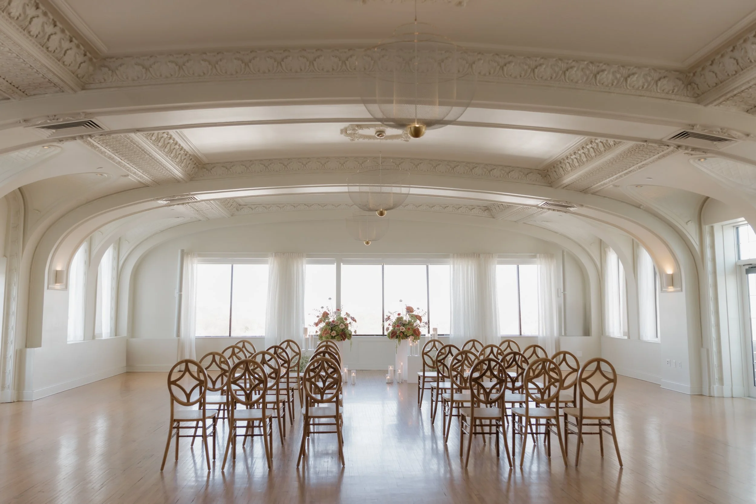 Empty event hall with white walls, ornate ceiling, wooden chairs arranged in a semi-circle, floral arrangements, and large windows with white curtains.