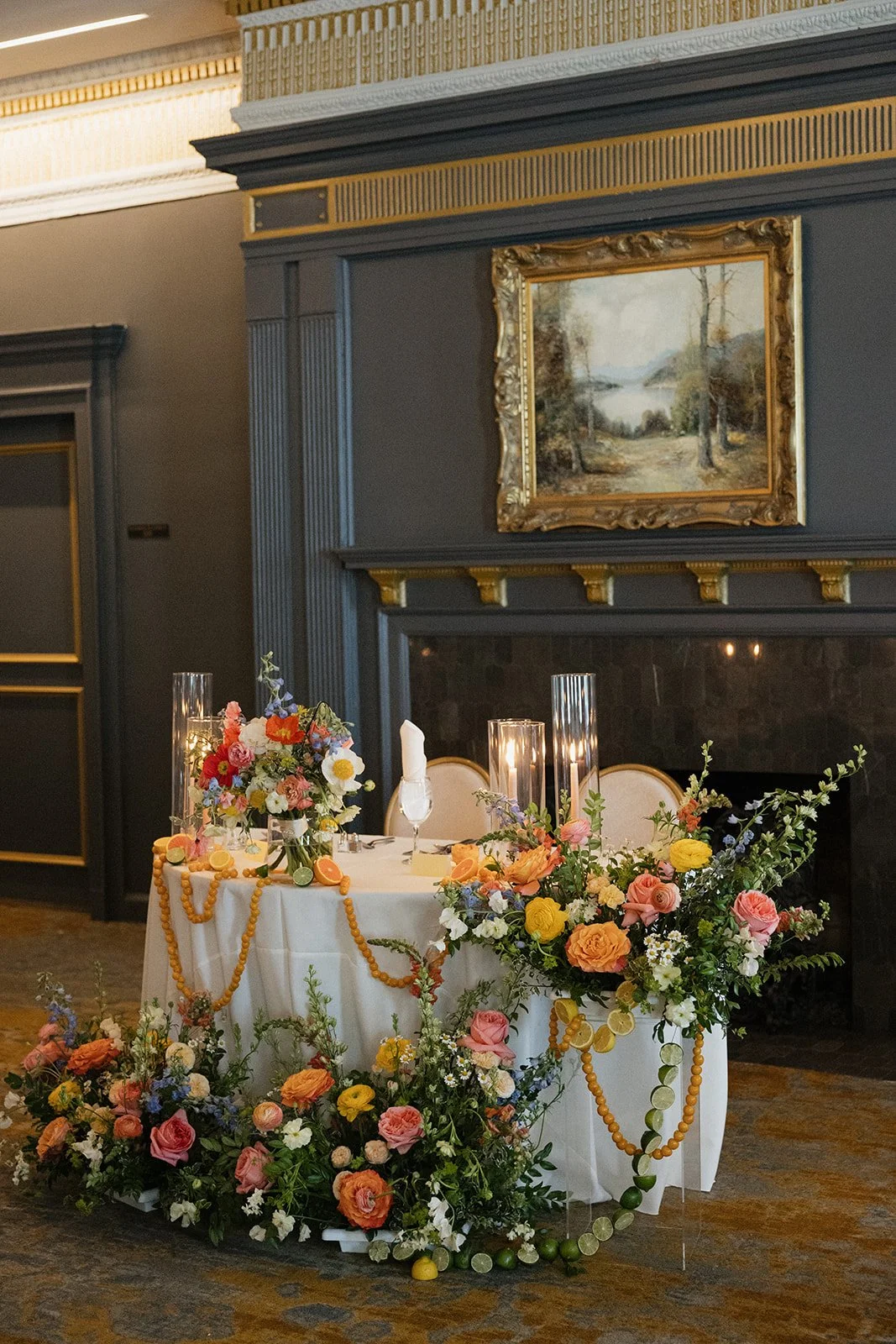 Decorated table with floral arrangements, candles, and citrus fruit slices in a formal room with dark walls, gold accents, a fireplace, and a landscape painting.