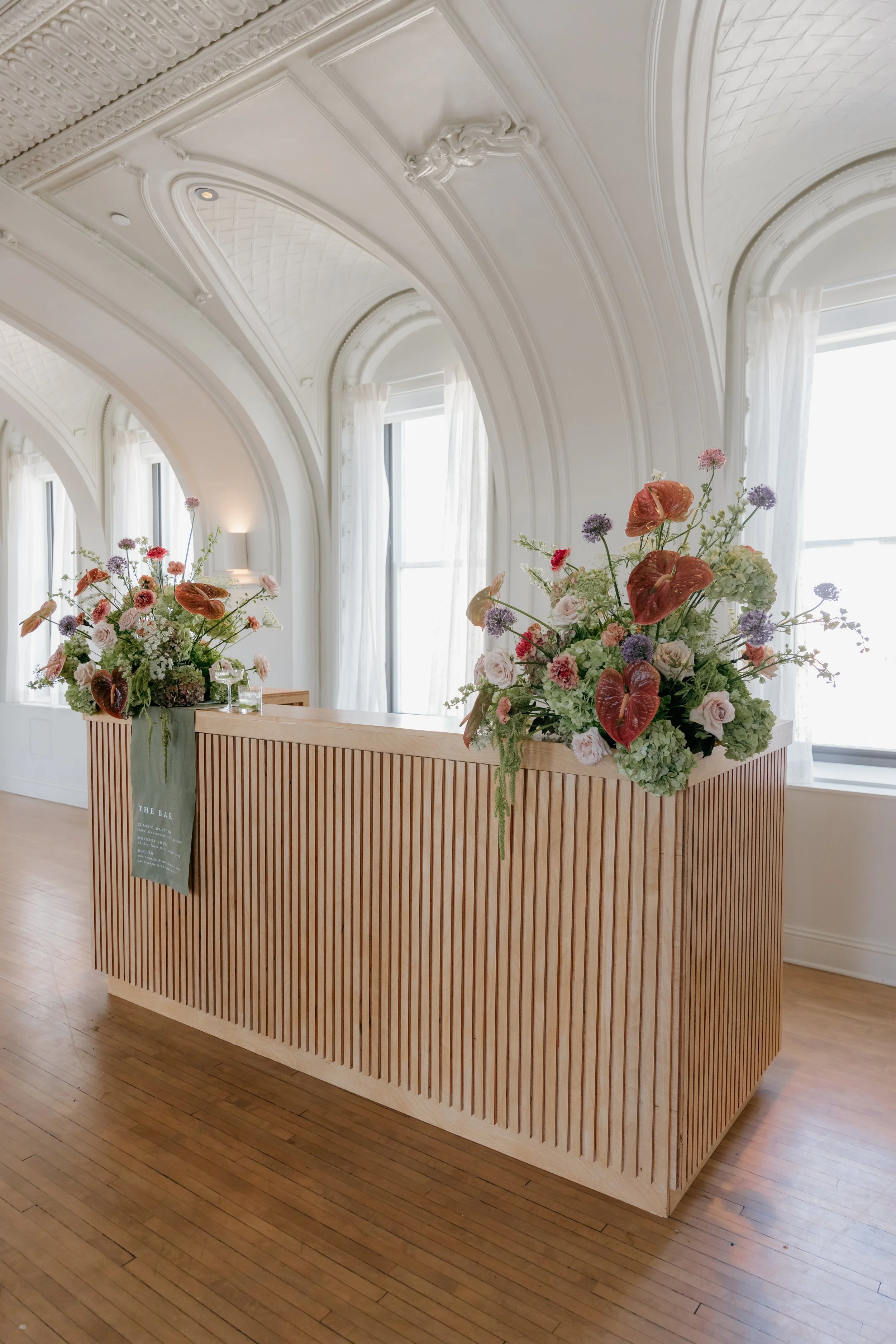 Elegant reception area with a wooden slatted counter topped with large floral arrangements featuring pink roses, green hydrangeas, purple alliums, and red anthuriums, set in a bright room with ornate white ceilings and large windows with sheer curtai