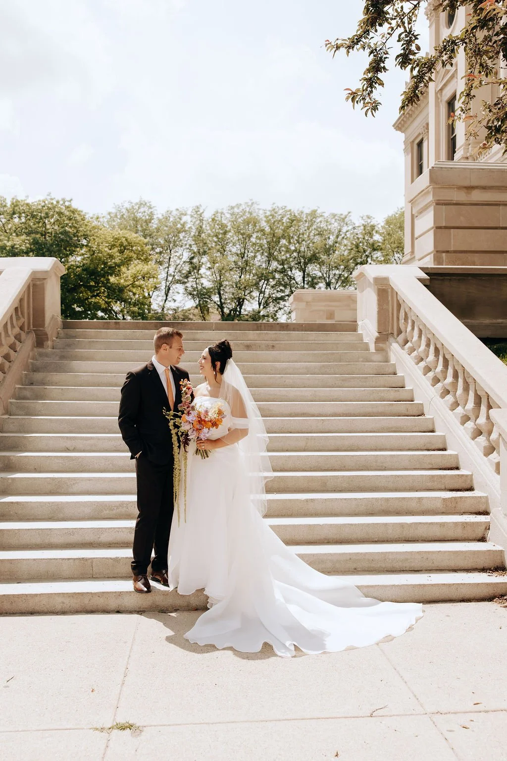 Bride and groom standing on outdoor staircase, facing each other, bride holding a bouquet of flowers, bride wearing a white wedding dress with veil, groom in black suit with tie.