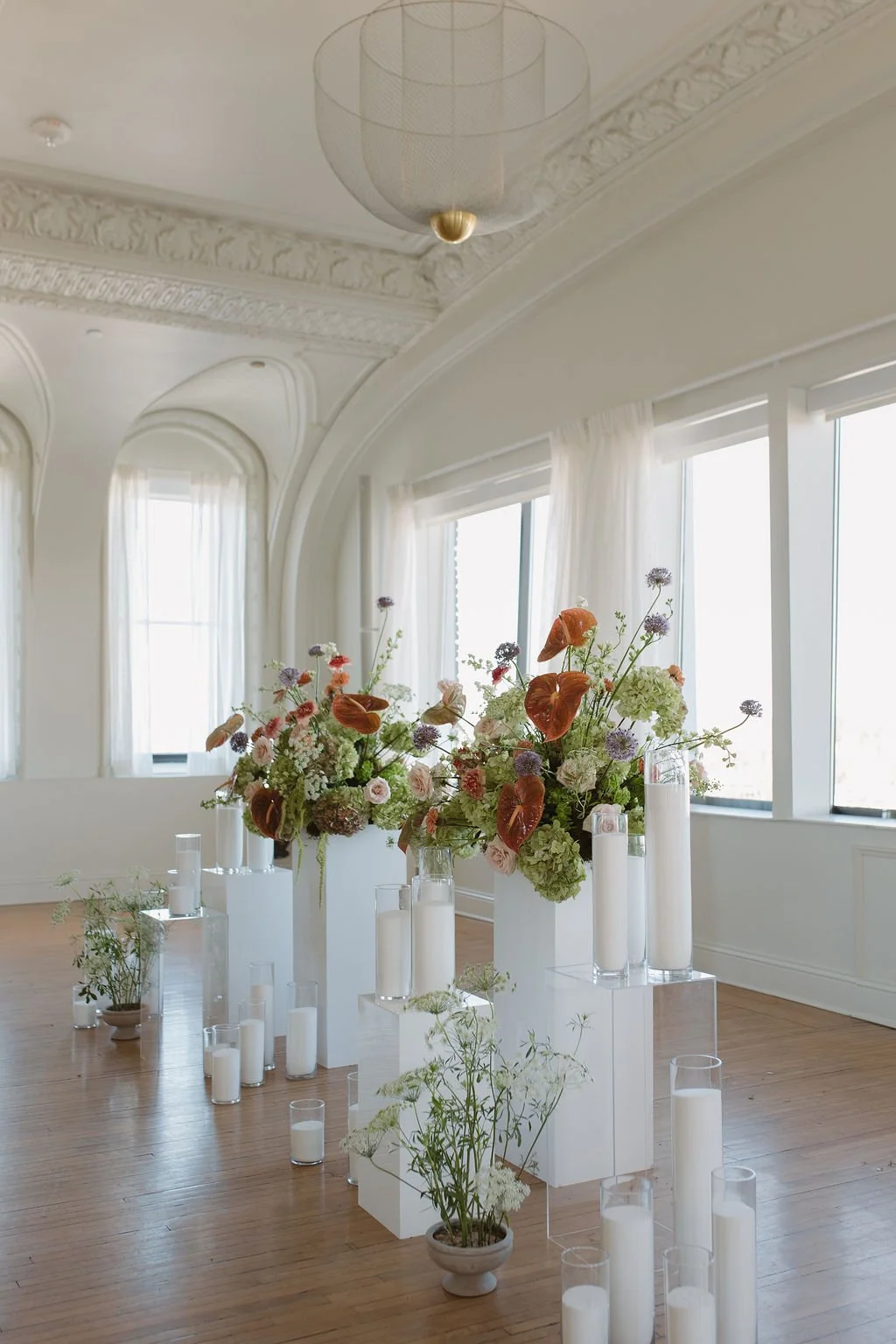 Elegant indoor space decorated with floral arrangements and candles for event, featuring large windows, white walls, and intricate ceiling moldings.