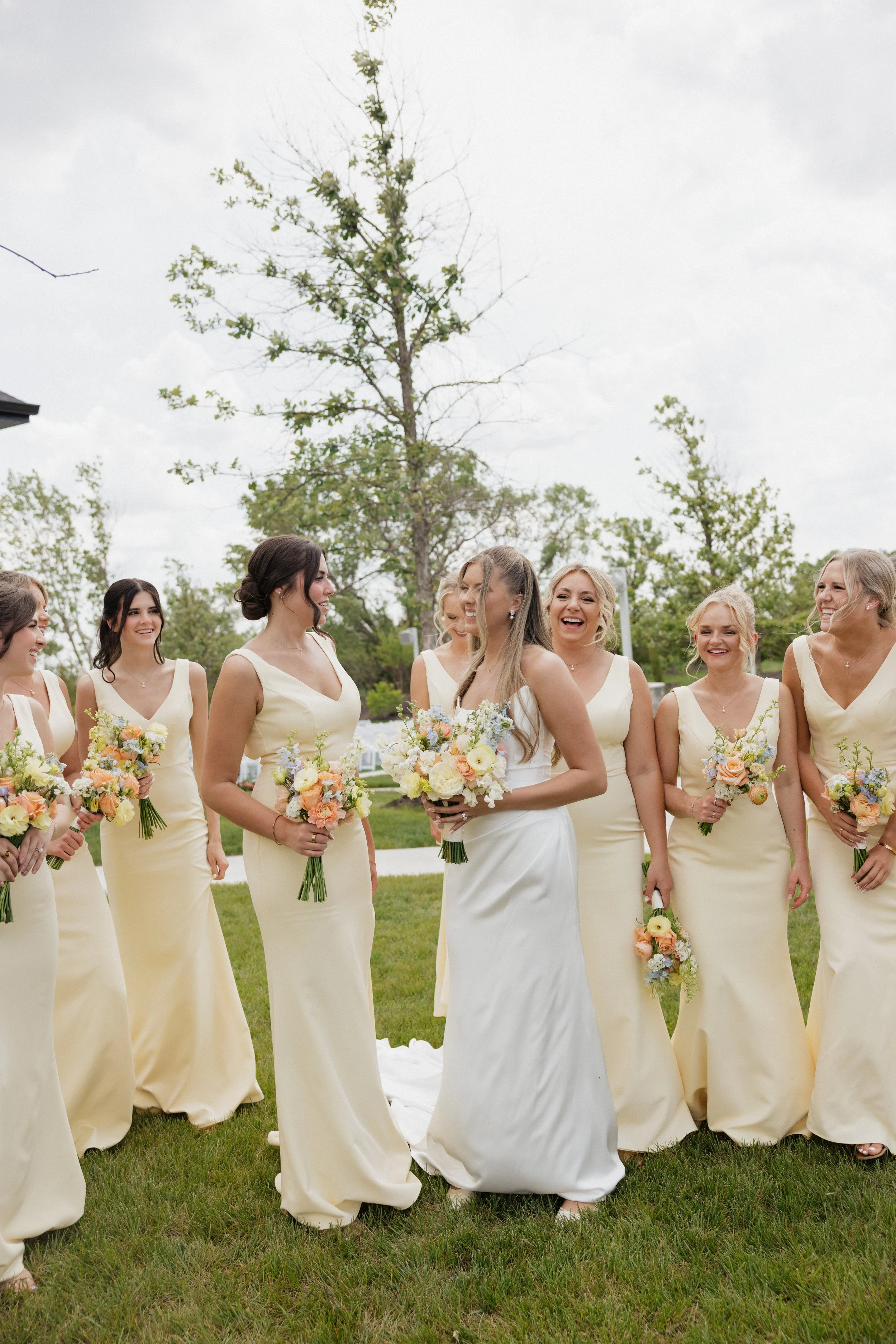 A group of bridesmaids in light yellow dresses are gathered outdoors on grass, holding bouquets of flowers, smiling and laughing at a wedding.
