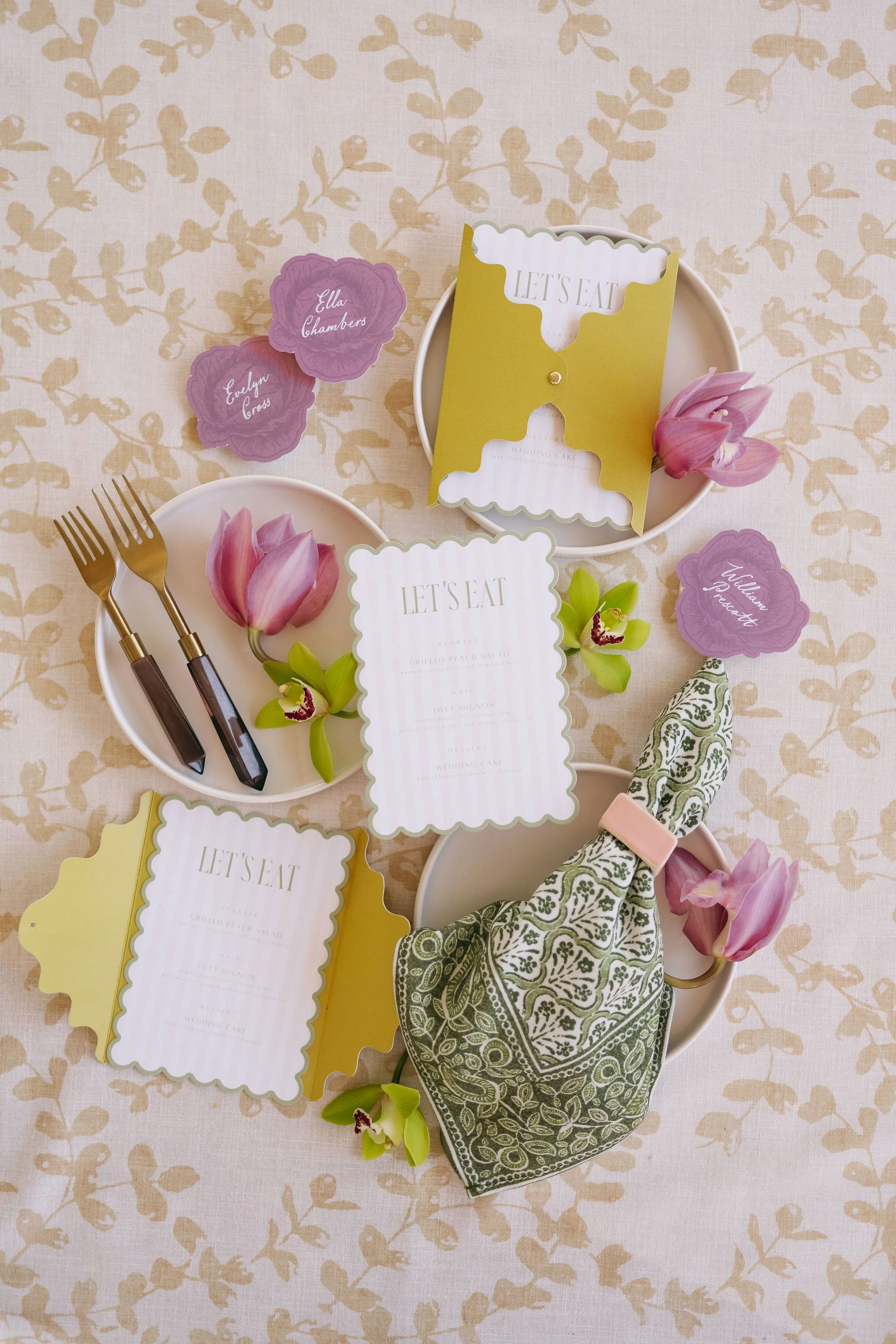 Table setting with wedding theme, including plates, gold forks, pink flowers, green patterned napkin, and wedding menu cards.