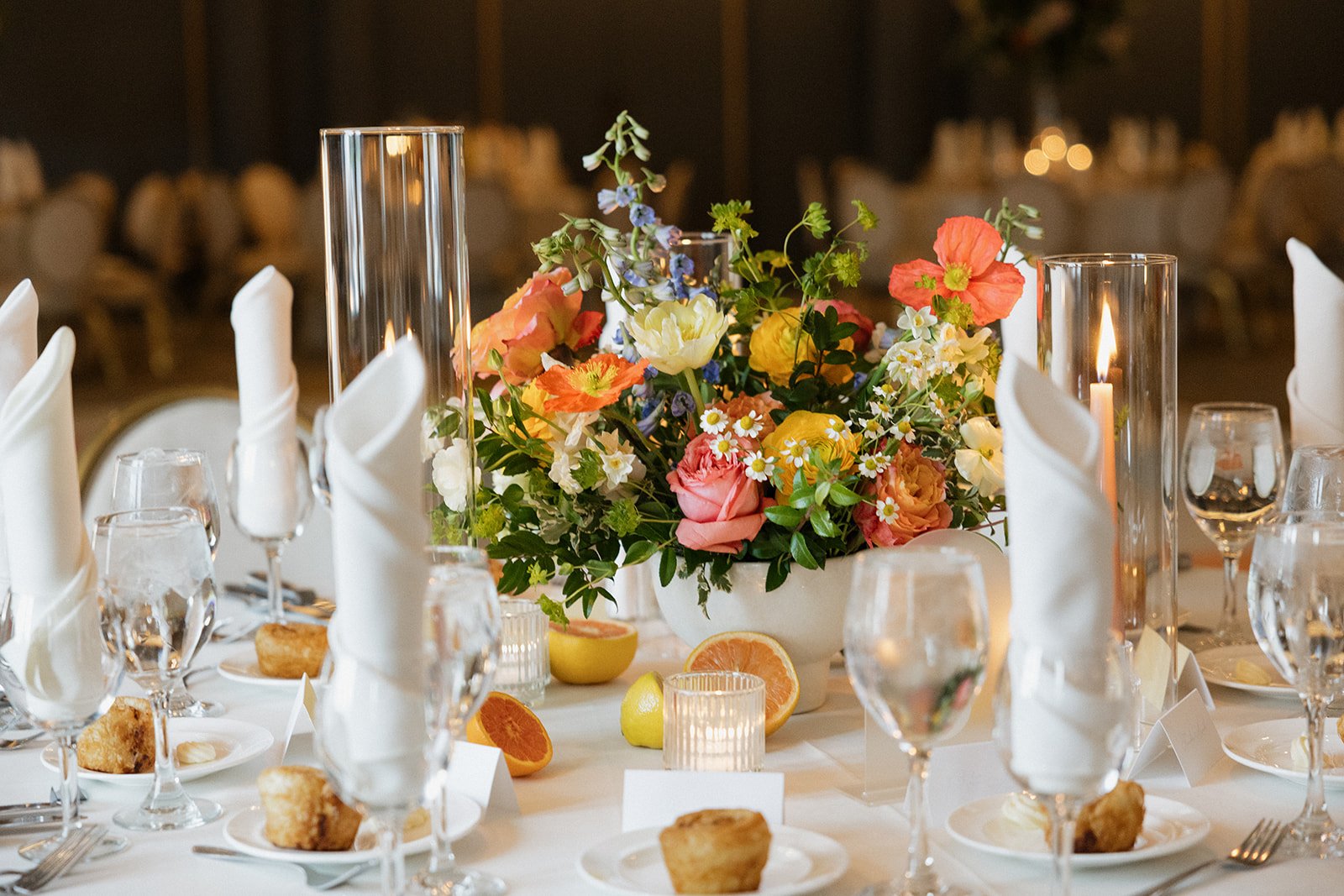 Elegant banquet table decorated with a large colorful floral centerpiece, surrounded by champagne glasses, water glasses, white napkins folded into cones, and plates with bread, in a dimly lit banquet hall.