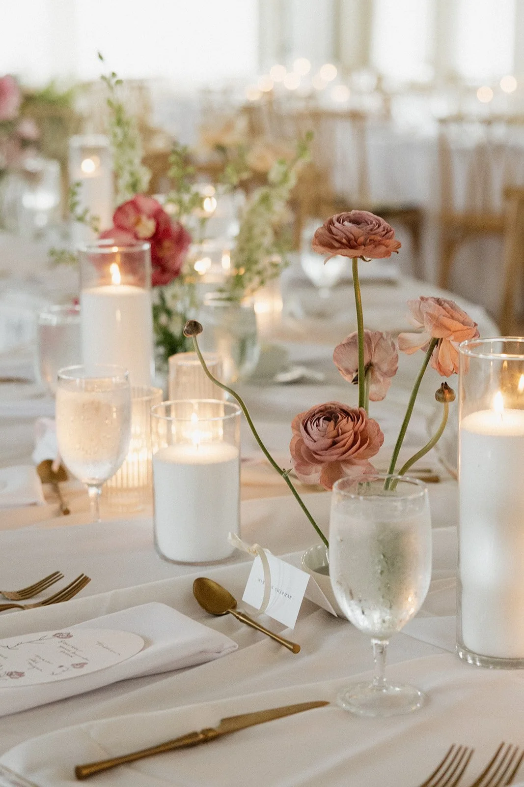 Elegant dining table decorated with pink ranunculus flowers in a vase, tall white candles in glass holders, and gold cutlery.