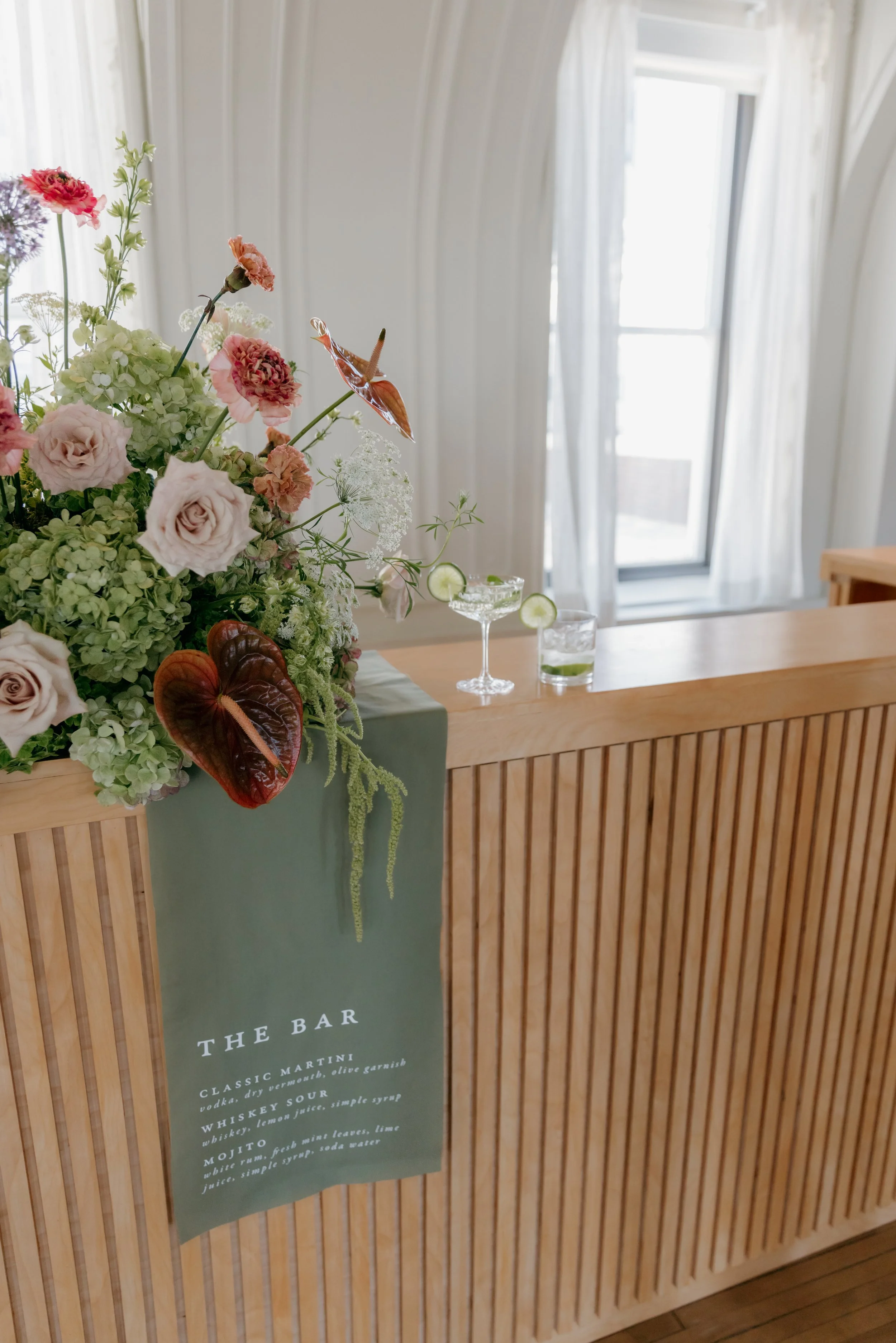 A wooden bar counter with a green banner that reads 'The Bar' and lists drinks. There is a large floral arrangement with roses, hydrangeas, and anthuriums on the left, and two cocktails with cucumber slices and a glass of lime water on the right. Bri