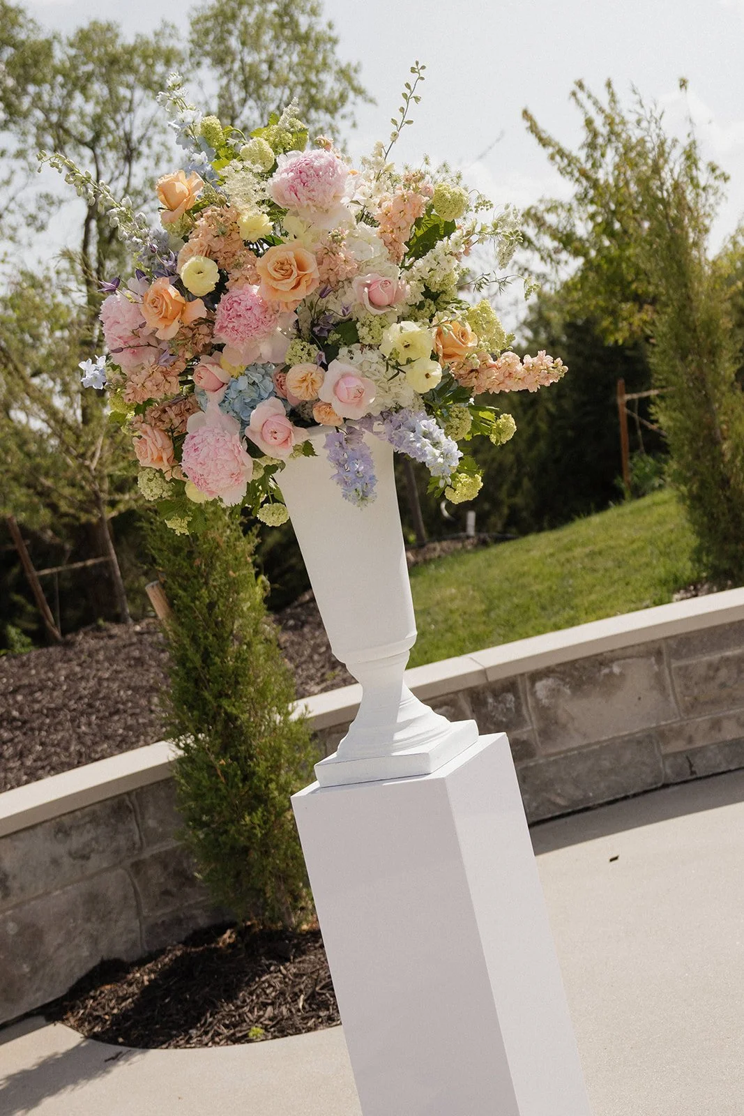 A large colorful flower arrangement in a white vase on a pedestal outdoors, with trees and a grassy area in the background.