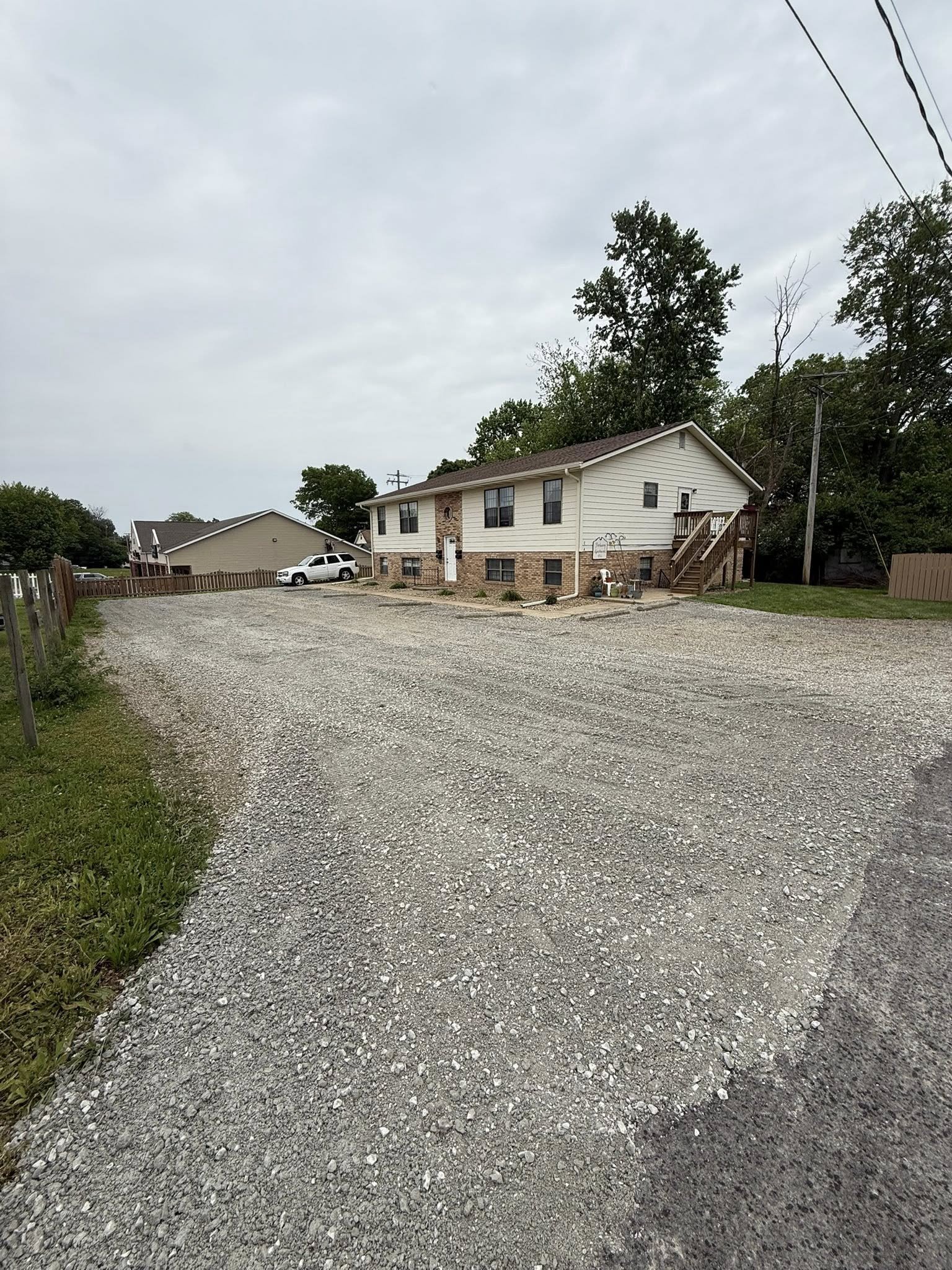 A gravel driveway in front of a house with white siding and brick foundation. There is a small wooden staircase on the right side leading to the back door. A white vehicle is parked near the house, and a fenced yard is visible in the background with 