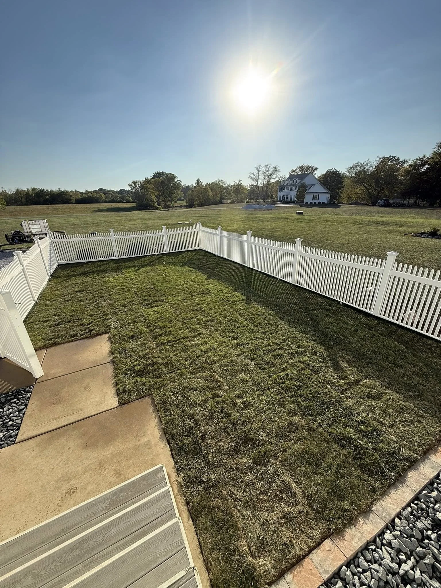Backyard with a white picket fence, green grass, a concrete patio, and a view of a large open field with trees and a house in the distance under a bright sun.