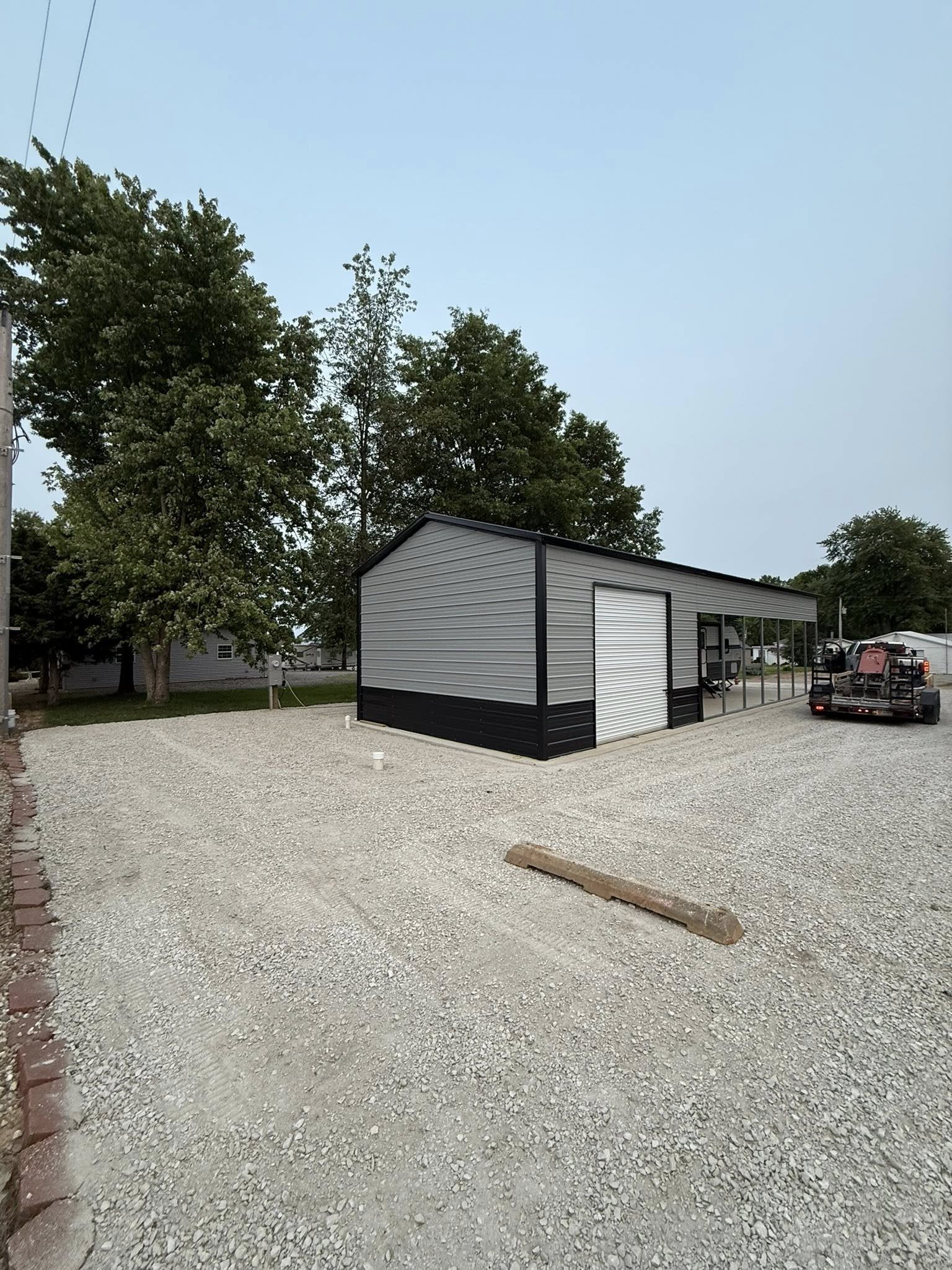 A gravel parking lot with a small metal storage building with a roller door and a side door. There are large trees in the background and a few trailers and vehicles parked nearby.