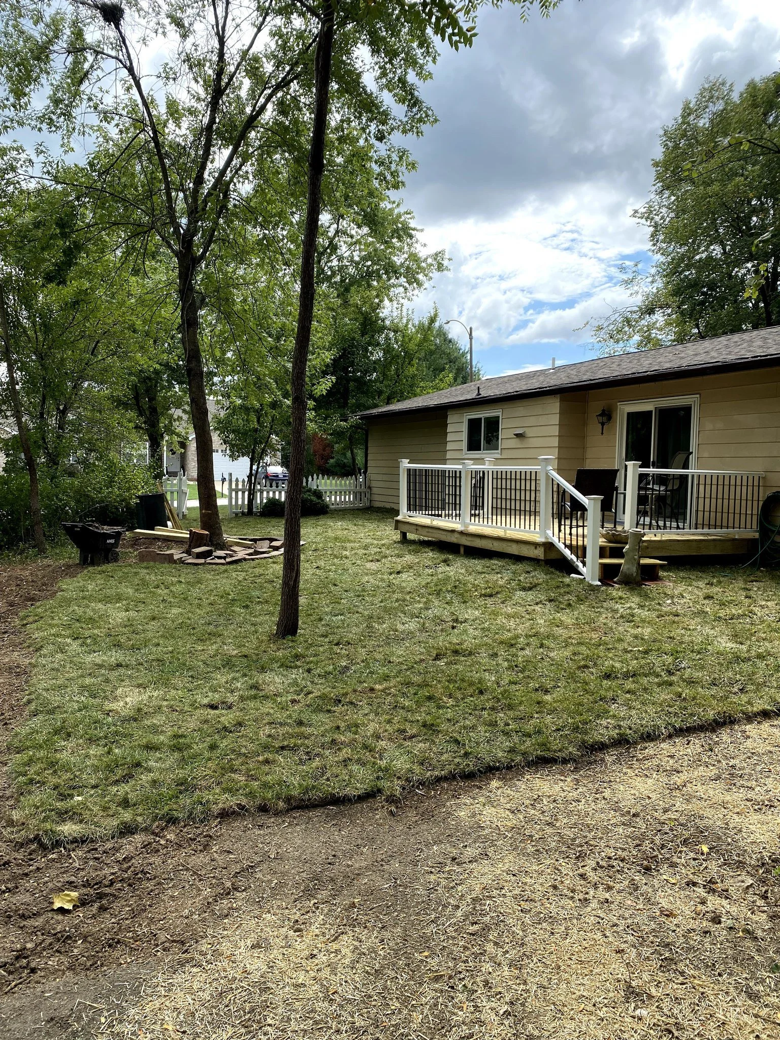 A backyard with a single-story house, a wooden deck, trees, and a grassy yard with some patches of dirt, under a partly cloudy sky.