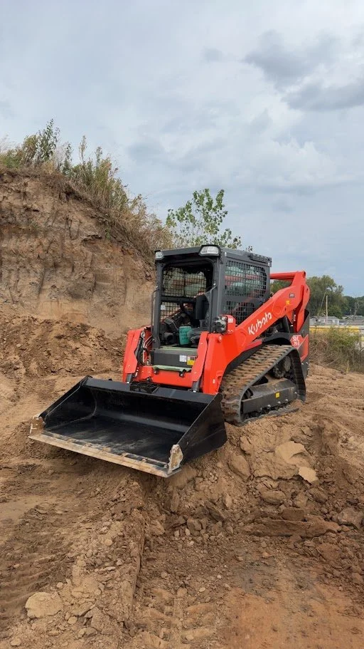 A Kubota compact track loader in orange and black, excavating dirt on a construction site with a dirt hill and cloudy sky in the background.