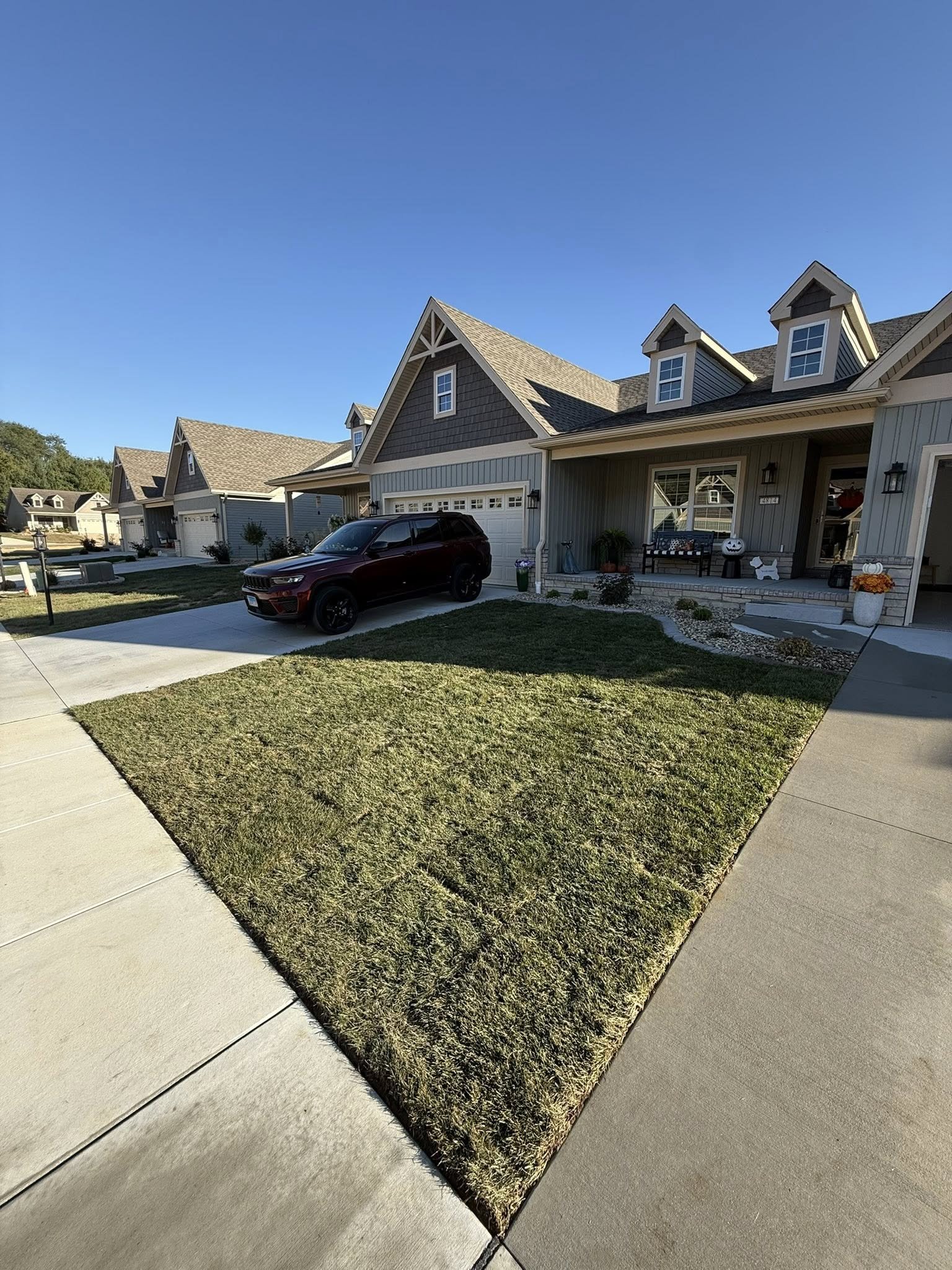 A suburban neighborhood with a row of houses, a driveway with a maroon SUV, a well-manicured lawn, and Halloween decorations including pumpkins and a scarecrow on the porch.