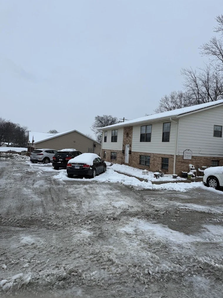 A snowy parking lot outside a two-story white and brick apartment building with several parked cars, some covered in snow, and leafless trees in the background.