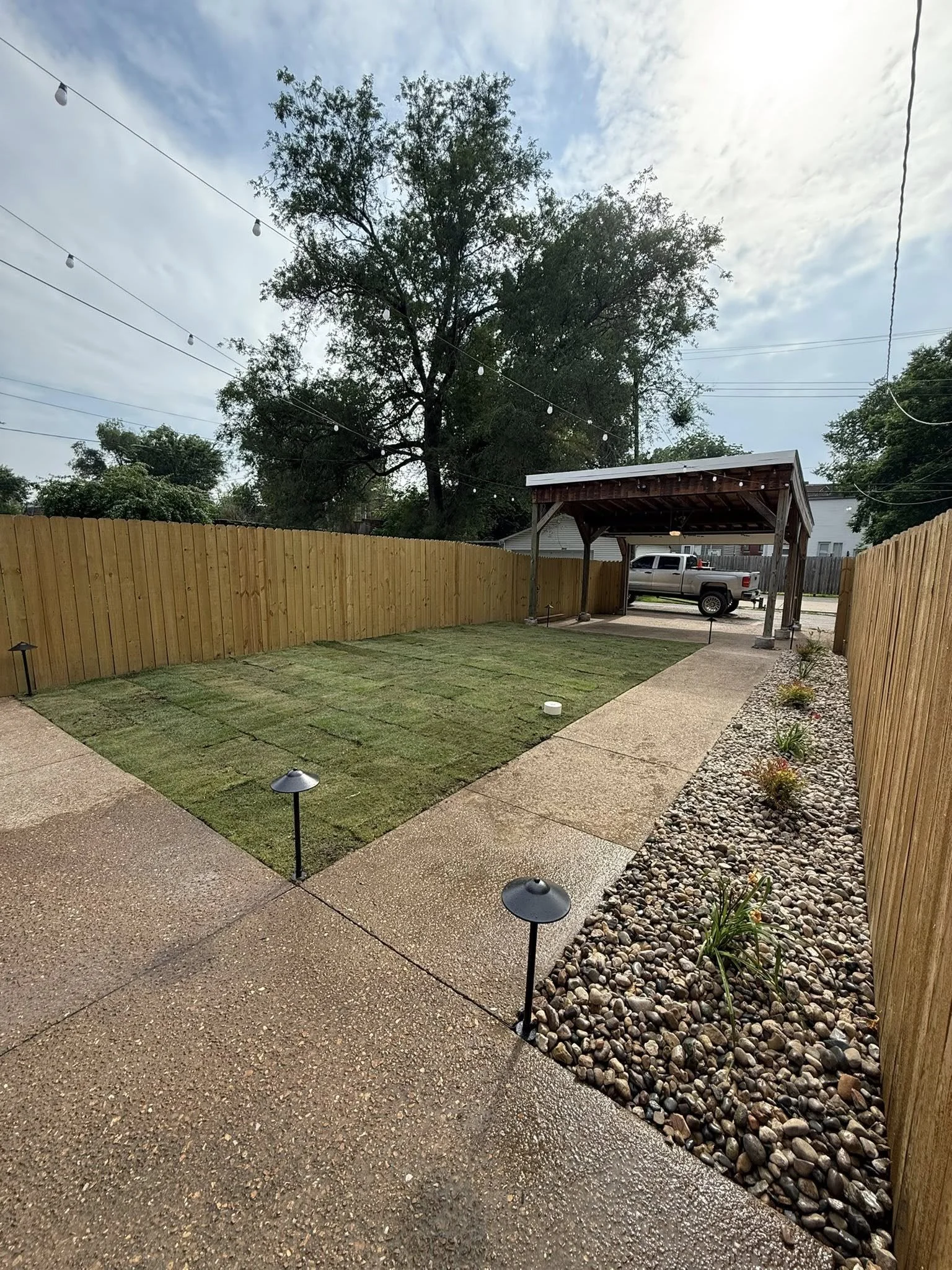 A backyard with a small grass patch, a concrete sidewalk, and a wooden fence. There are small lights along the sidewalk, a shed with a truck parked underneath, and a large tree in the background. The sky is partly cloudy.