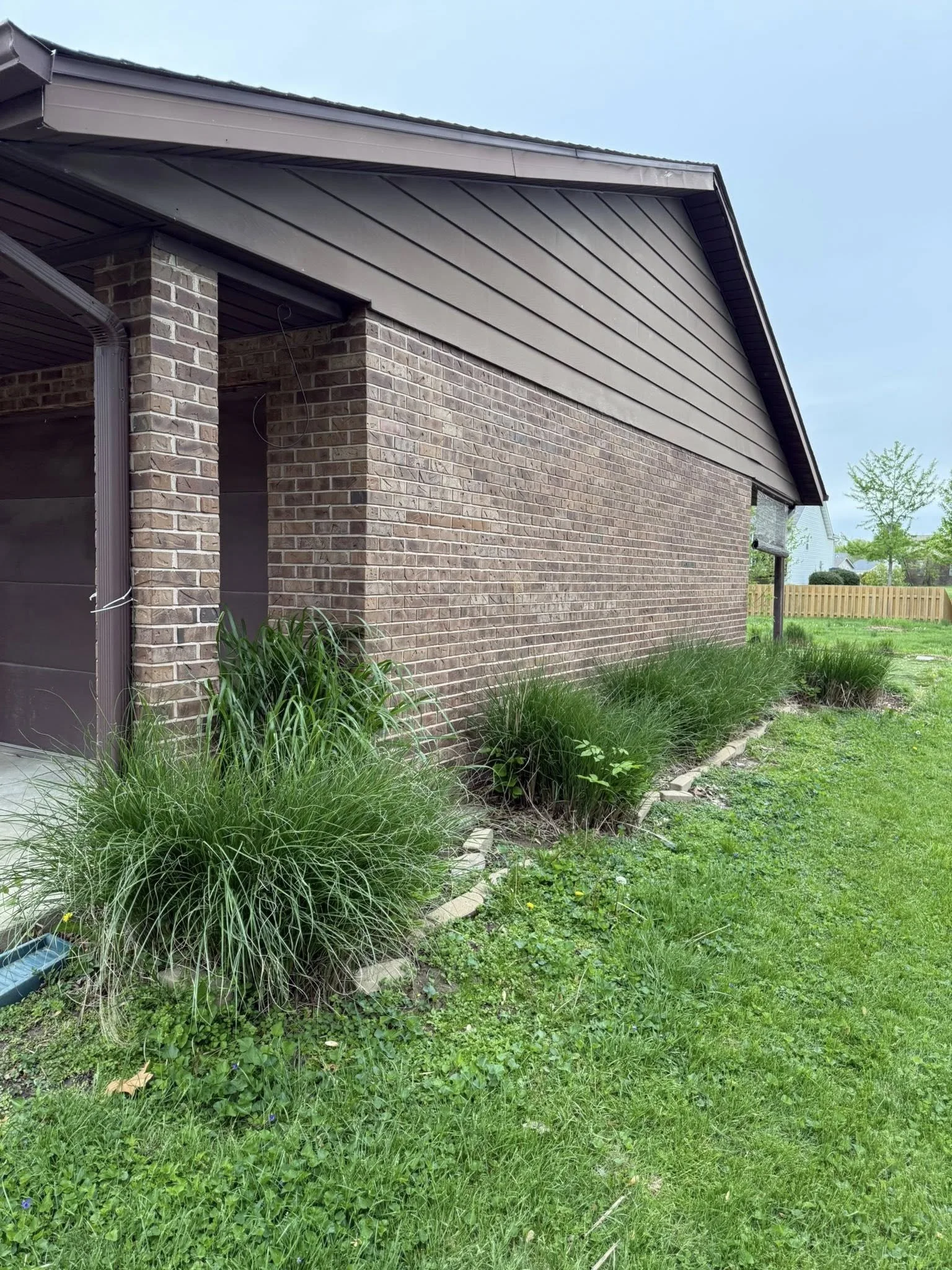 Side view of a brick house with a sloped roof, a small garden bed with green plants along the side, and a fenced backyard in the background.