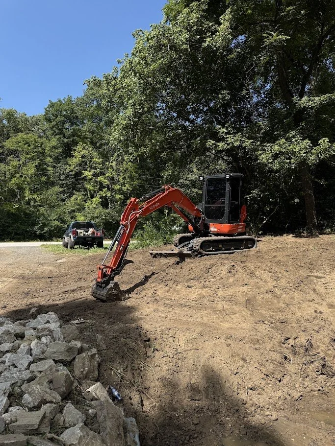 A small orange excavator digging into a dirt slope near a wooded area, with a black and silver pickup truck parked on the grassy ground in the background.