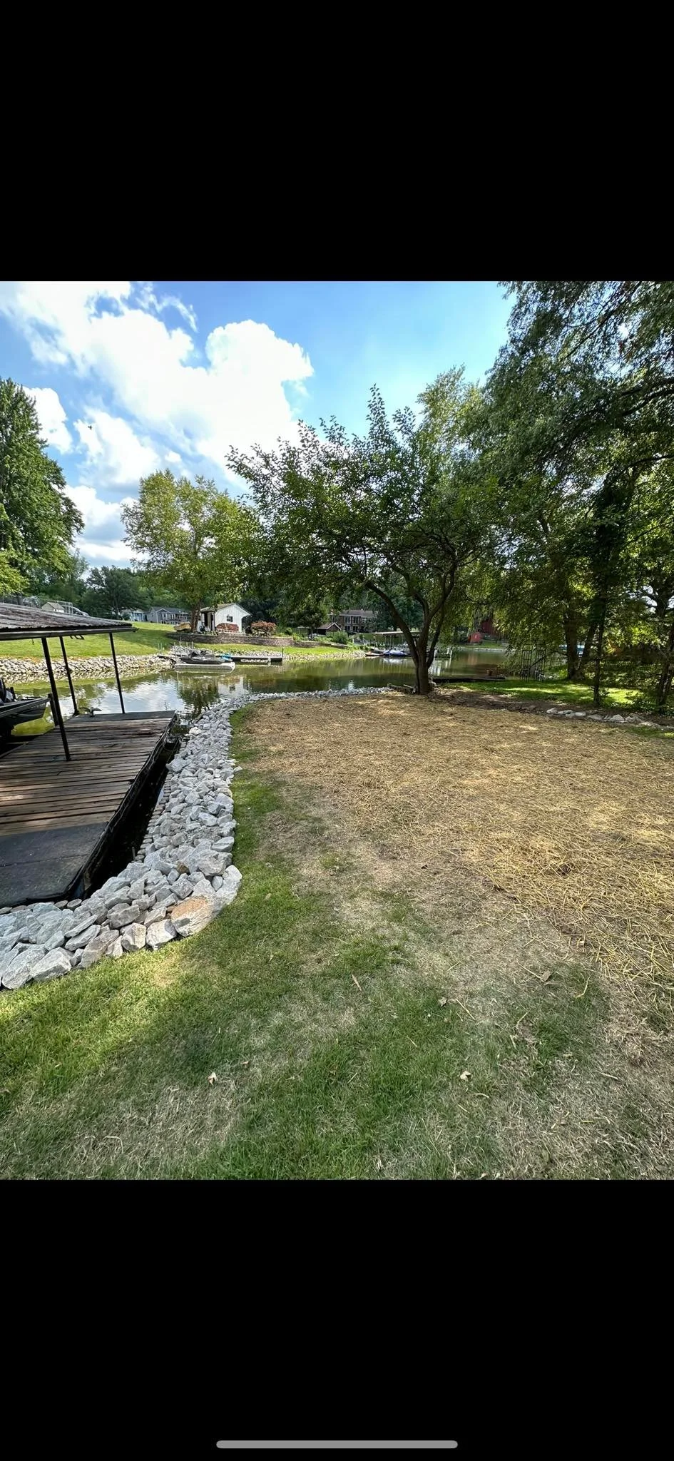 Lakeside backyard with a grassy area, a tree, boat docks, and boats on the water, under a partly cloudy sky.