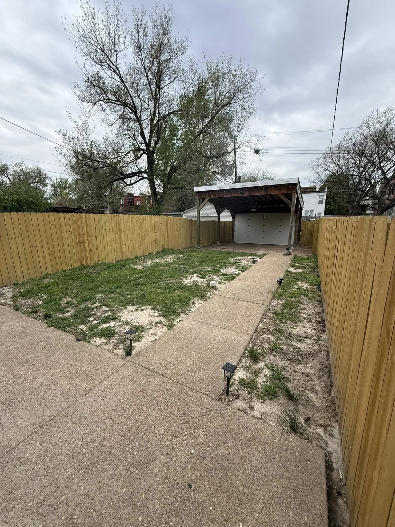 A backyard enclosed by a wooden fence with a concrete pathway leading to a small garage with an open door under a metal roof. The yard has a mix of grass and sandy patches with small solar lights along the pathway. Overcast sky with trees in the back