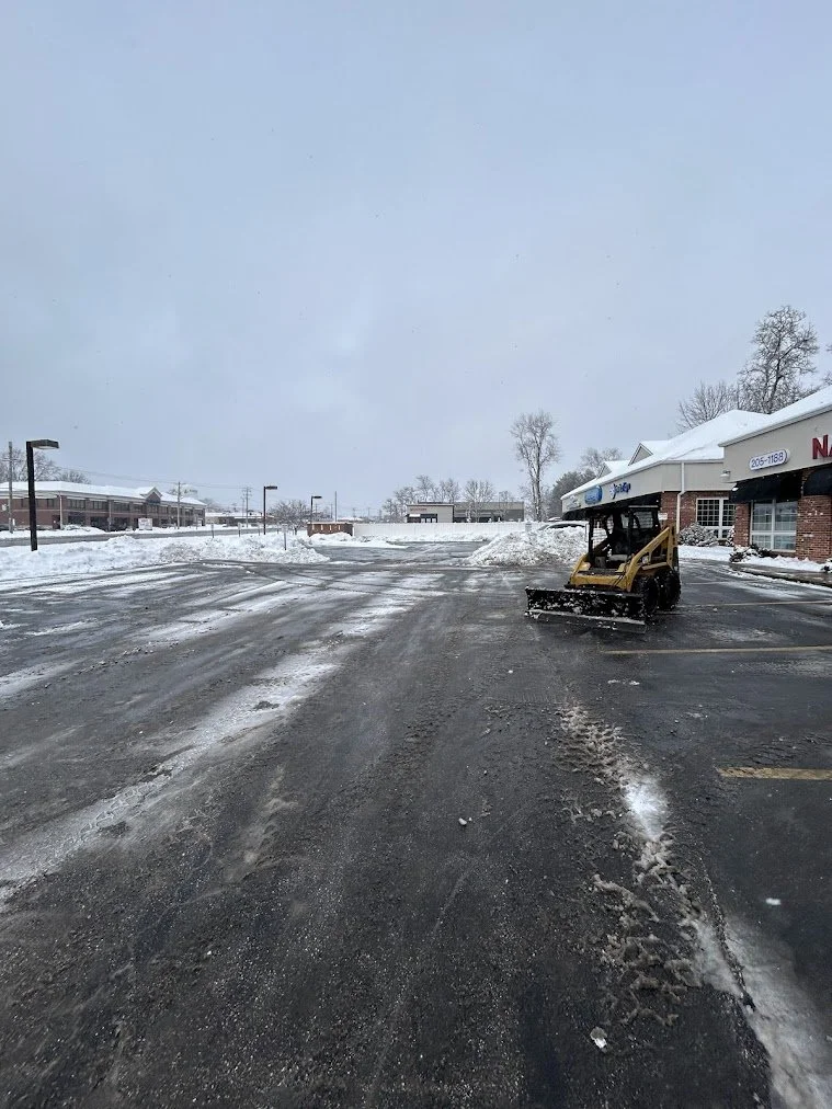 Empty parking lot covered in snow with a small snow removal vehicle parked near a shopping plaza in winter.