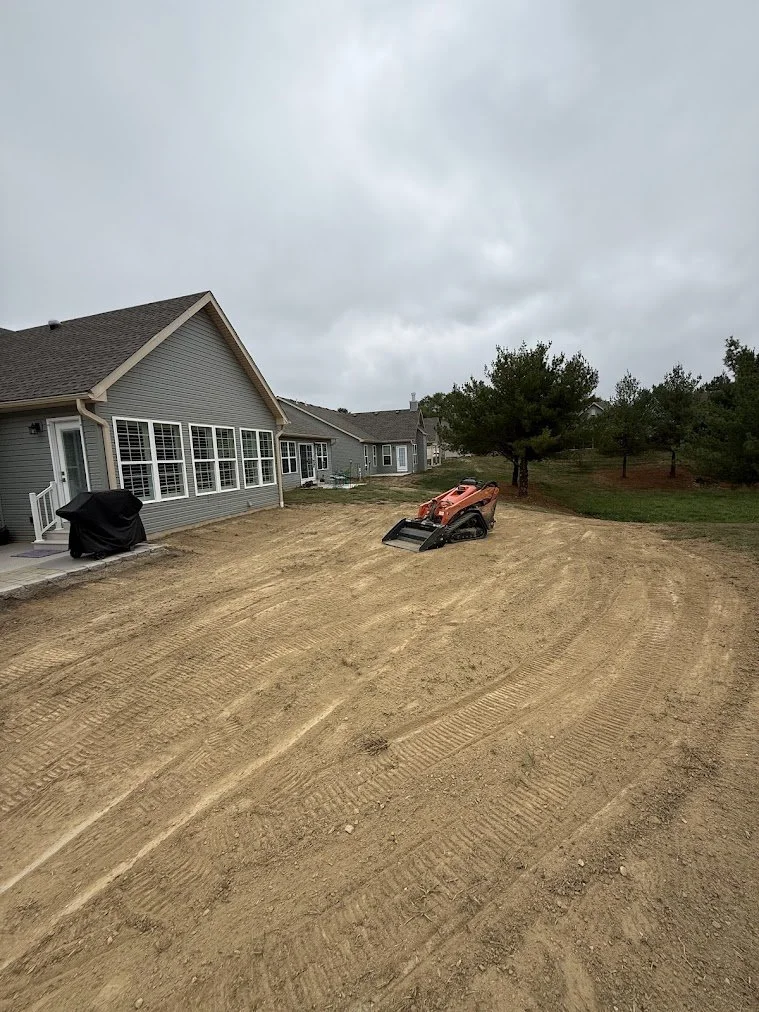 A backyard under construction with a dirt surface, a small orange compact excavator, and a gray house with multiple large windows and a covered barbecue grill.