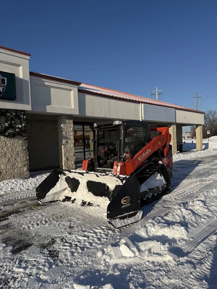 A Kubota skid steer loader with a snow blower attachment clearing snow outside a commercial building under a clear blue sky.
