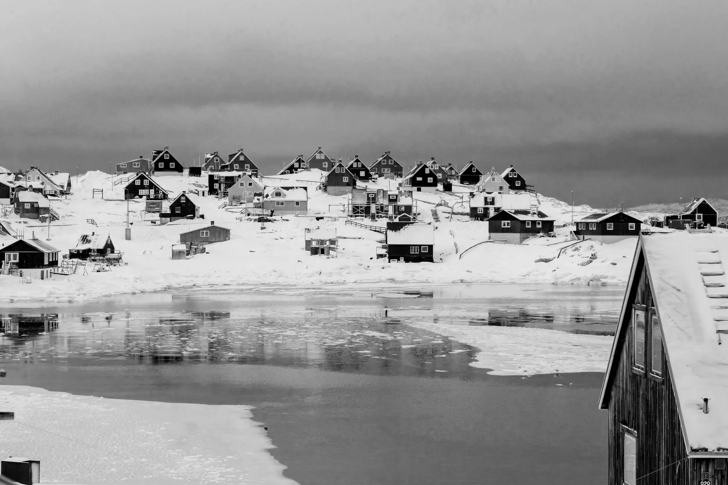 Snow-covered houses on a hillside near a partially frozen body of water under a cloudy sky.