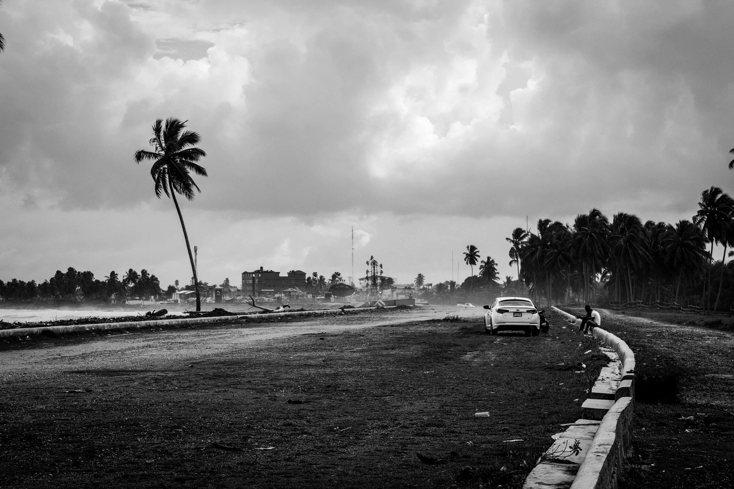 Black and white photo of a damaged road with a leaning palm tree, a car parked on the side, and a person sitting on the curb, with stormy clouds overhead and palm trees in the background.