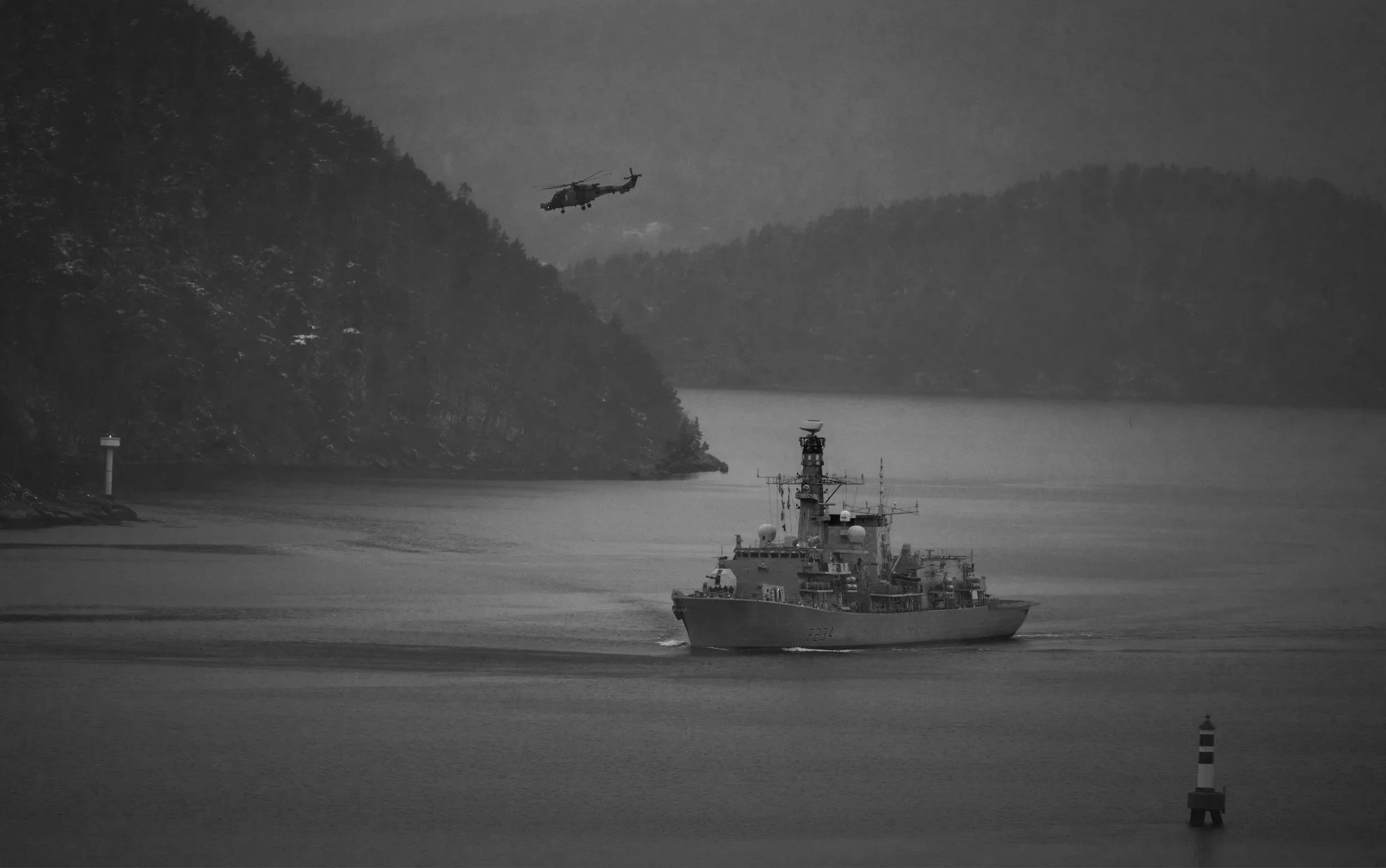 A navy ship sailing in the water with a helicopter flying overhead, near forested hills and a lighthouse at the water's edge.