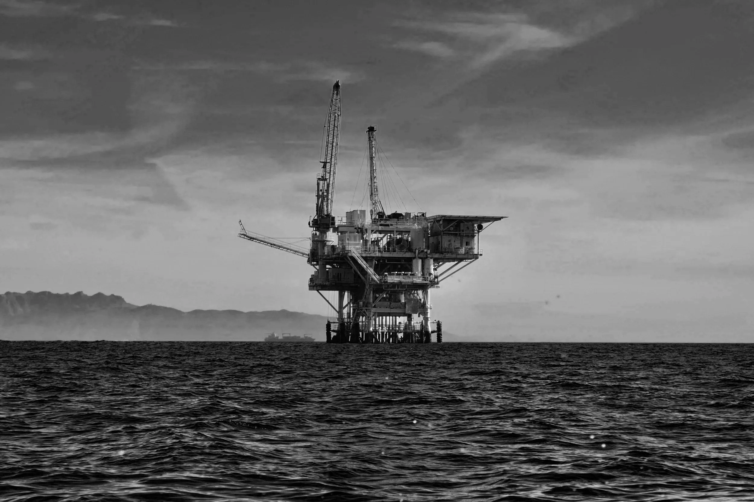 An offshore oil rig in the ocean, with a distant landmass in the background, in black and white.