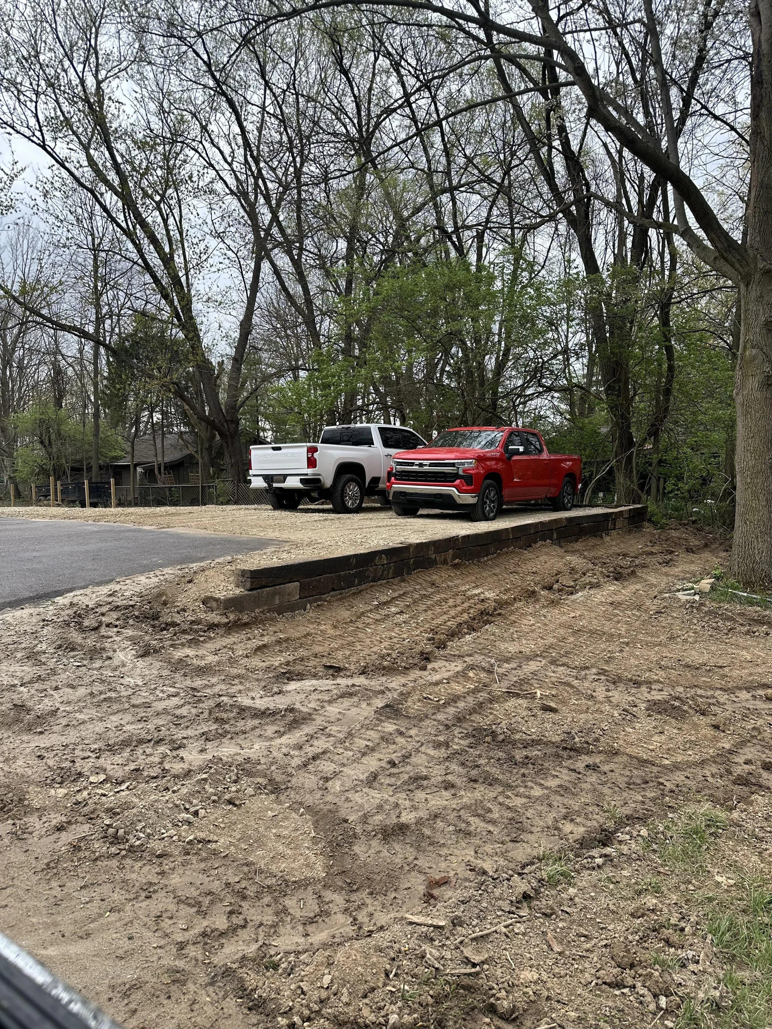 Two pickup trucks, one white and one red, parked on a dirt area next to a paved road with trees in the background.