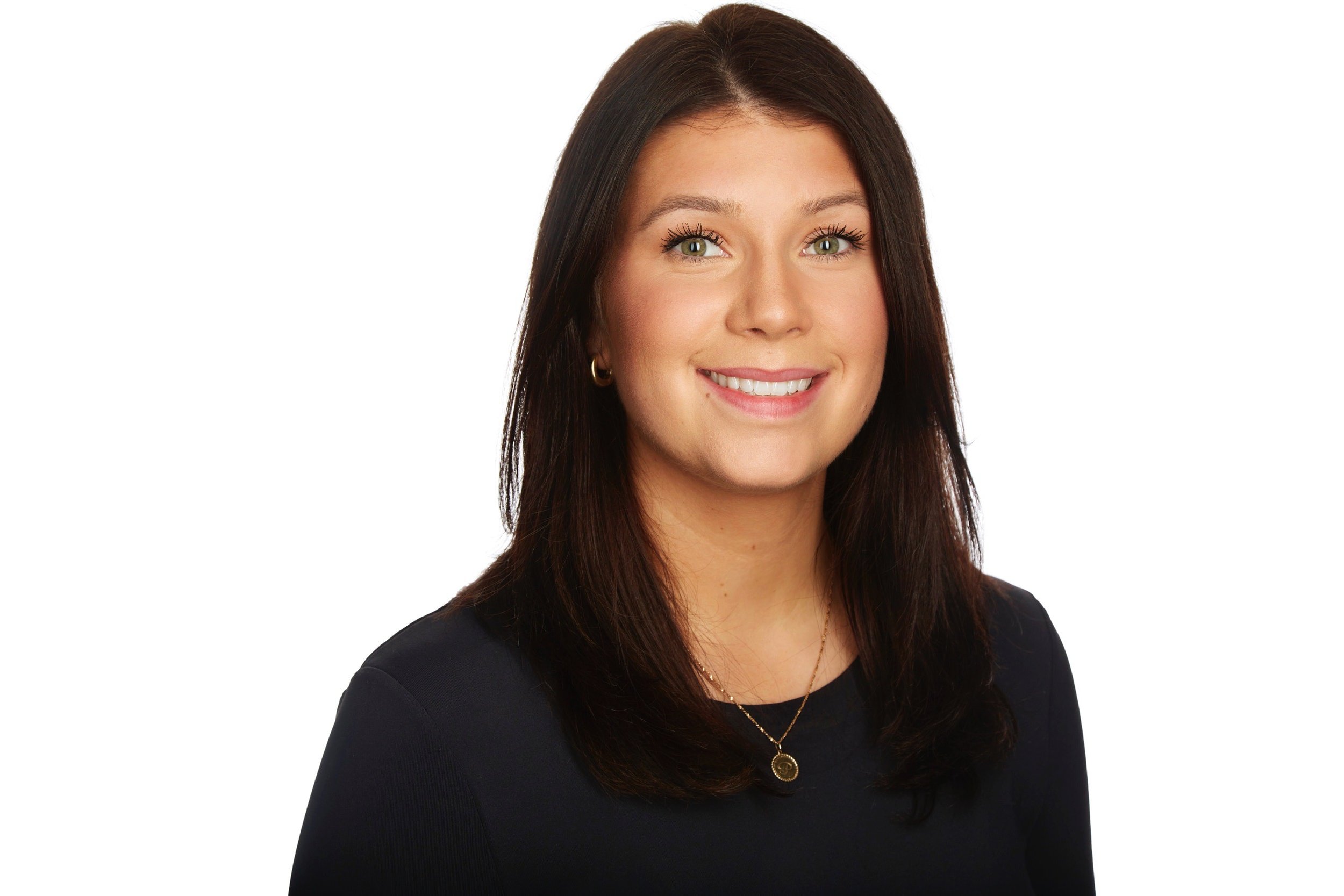 A woman with shoulder length dark brown hair and blue eyes, smiling, wearing a black top and a gold necklace with a pendant, against a white background.