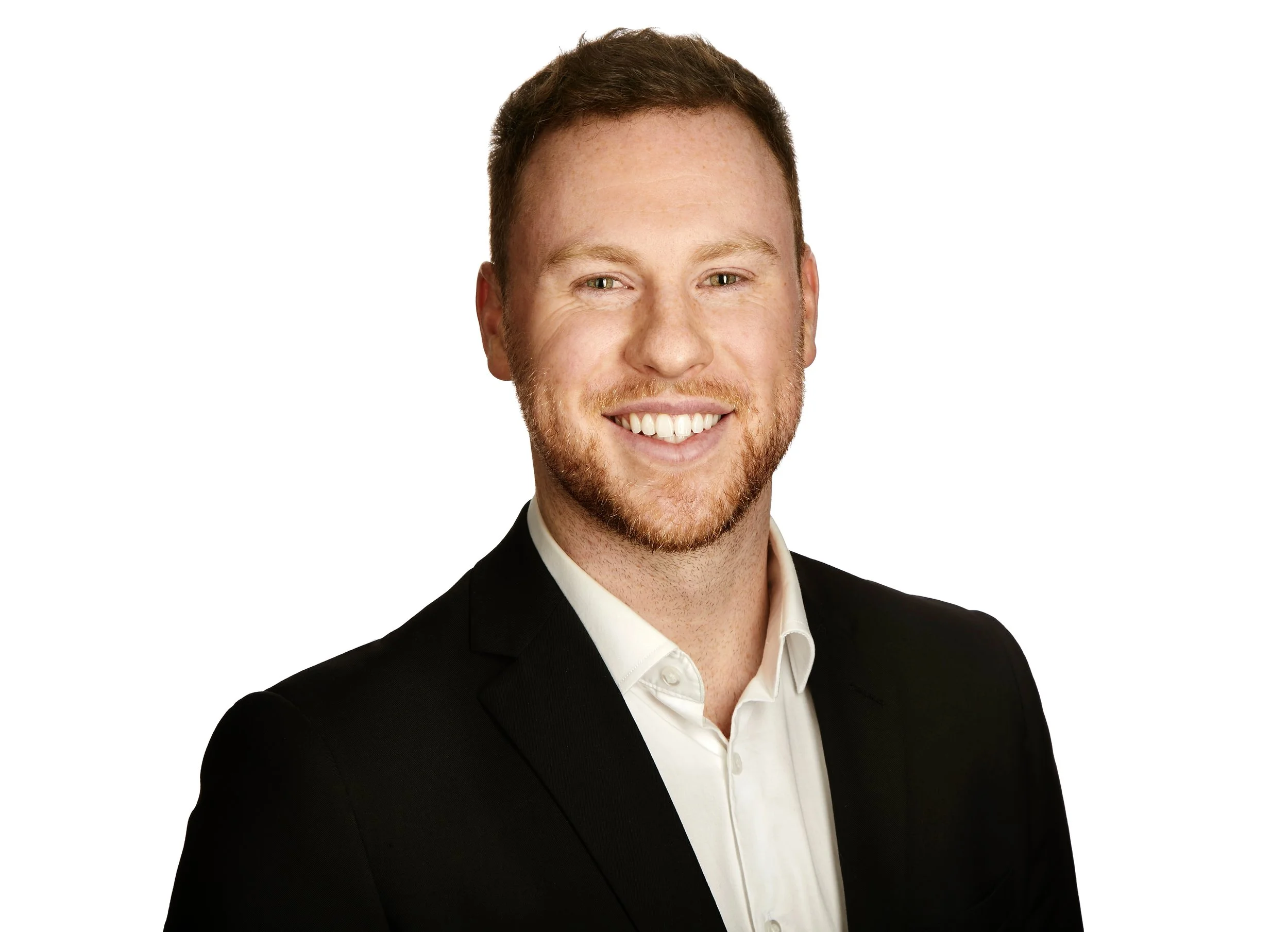 A smiling man with short reddish hair and a beard, dressed in a black suit and white shirt, photographed against a white background.